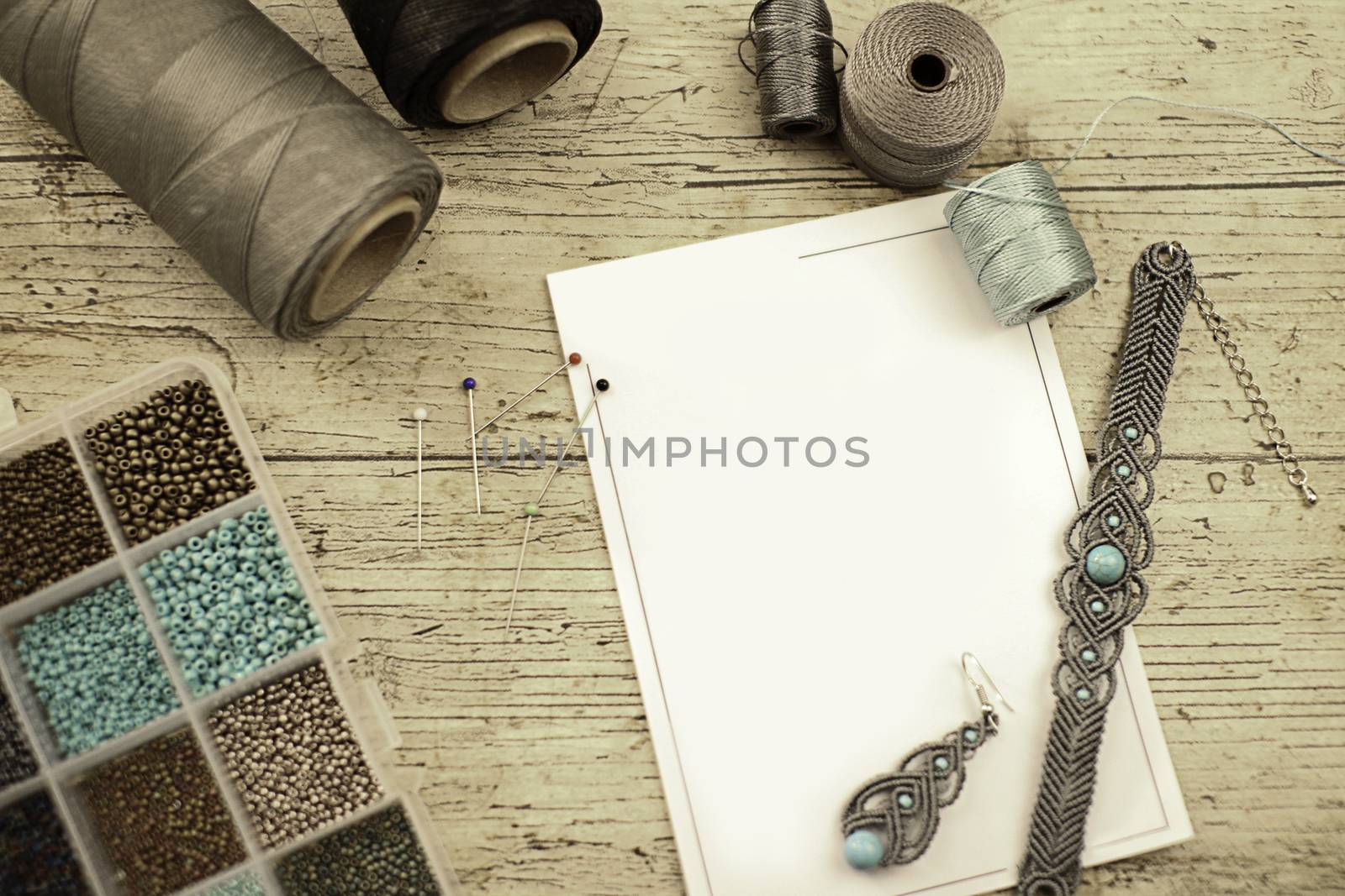 Top view of a wooden table with tools, spools of thread, natural stones and colored beads to make macramé jewelry, with blank sheet for copy space