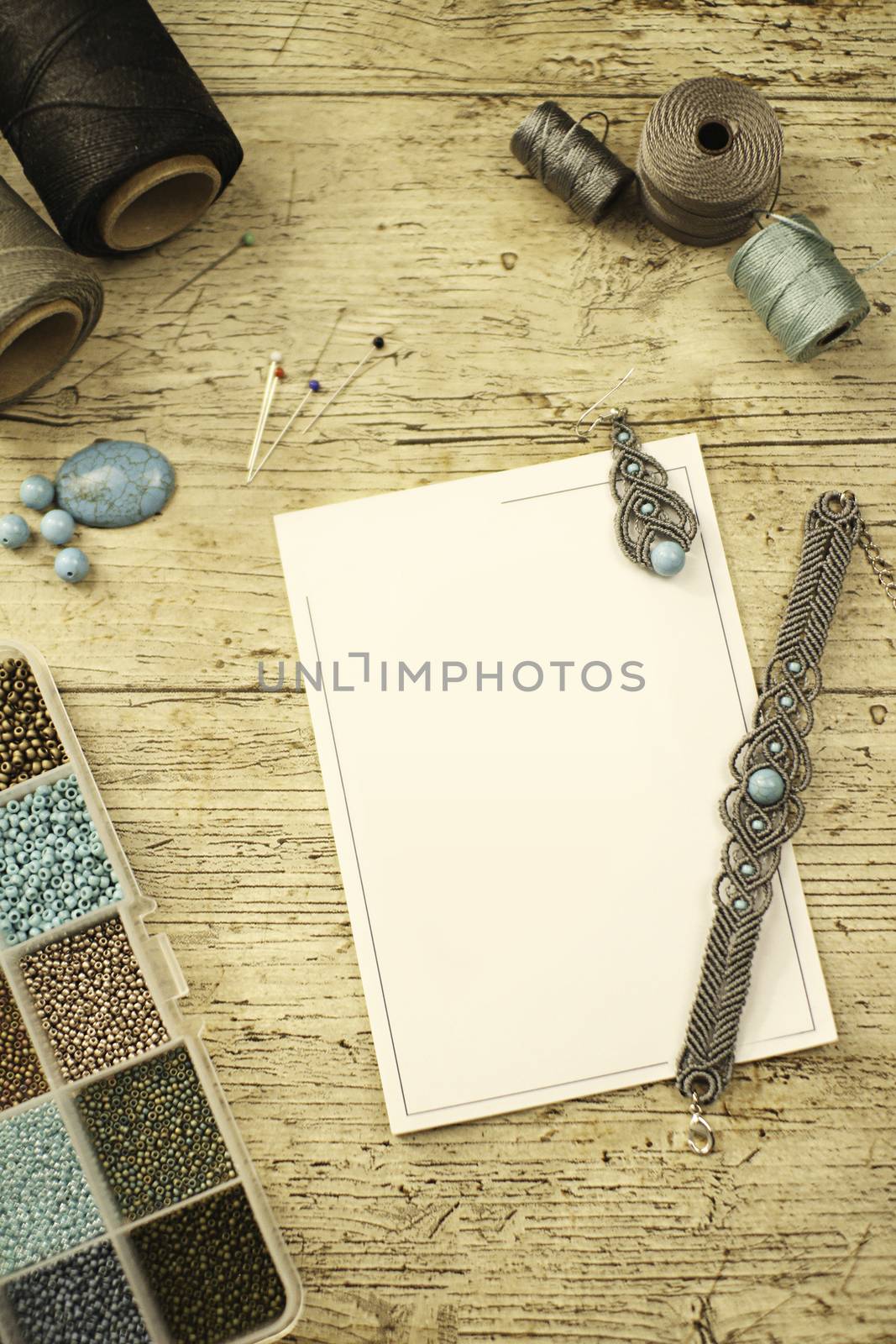 Top view of a wooden table with tools, spools of thread, natural stones and colored beads to make macramé jewelry, with blank sheet for copy space by robbyfontanesi