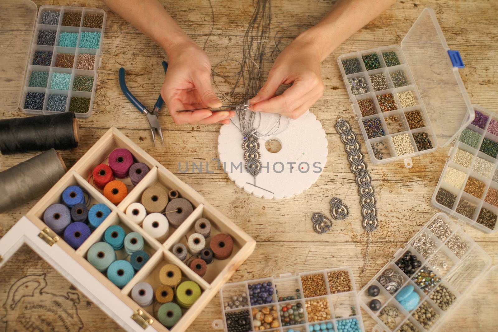 Top view of female hands making a macramé bracelet with kumihimo on a wooden table with tools, spools of thread, natural stones and colored beads by robbyfontanesi