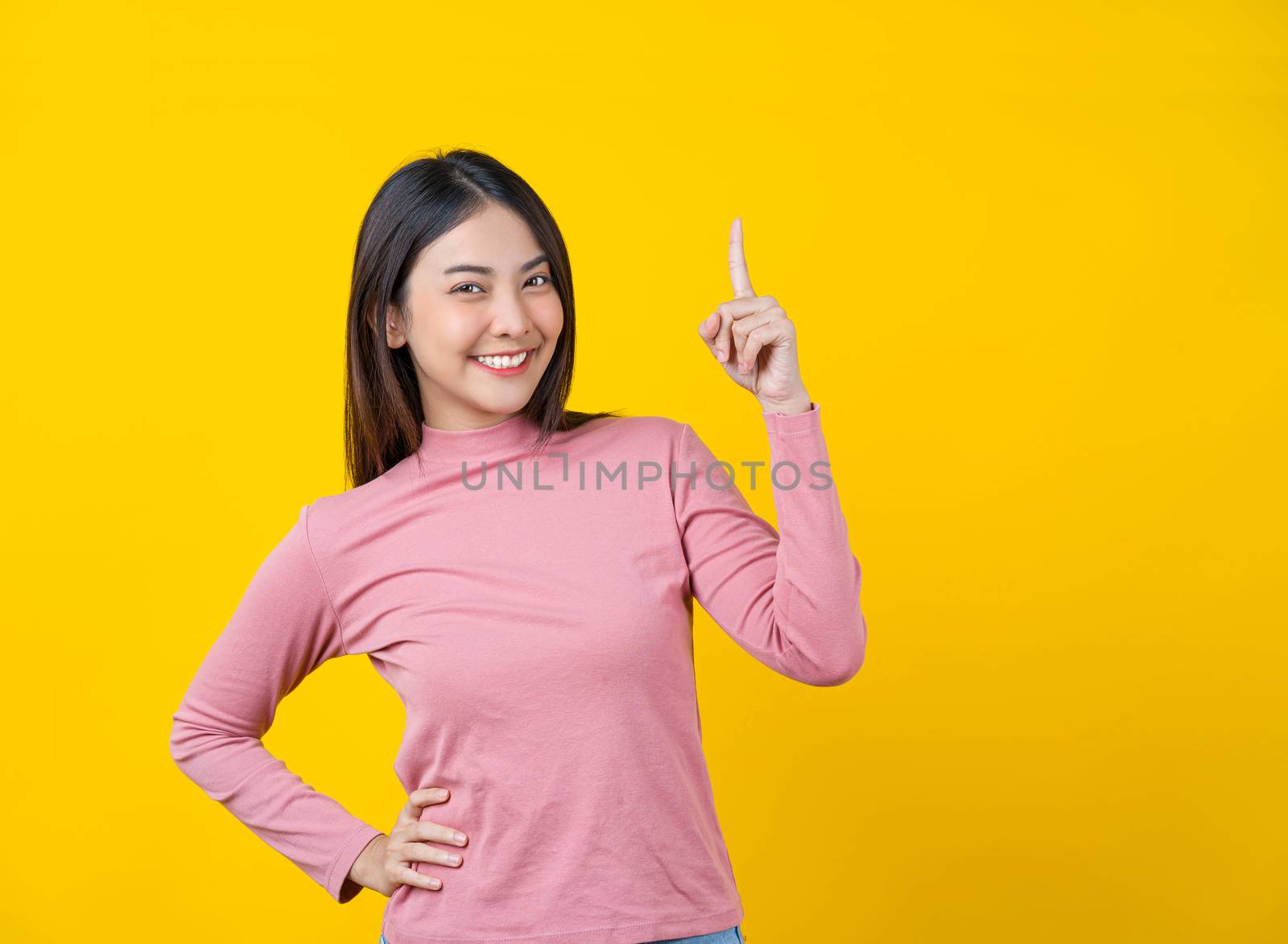 Asian smiling young woman finger Pointing up for advertise, banner or showing on isolated yellow color background, wearing winter cozy sweater indoors studio, happiness and fun, copy space concept