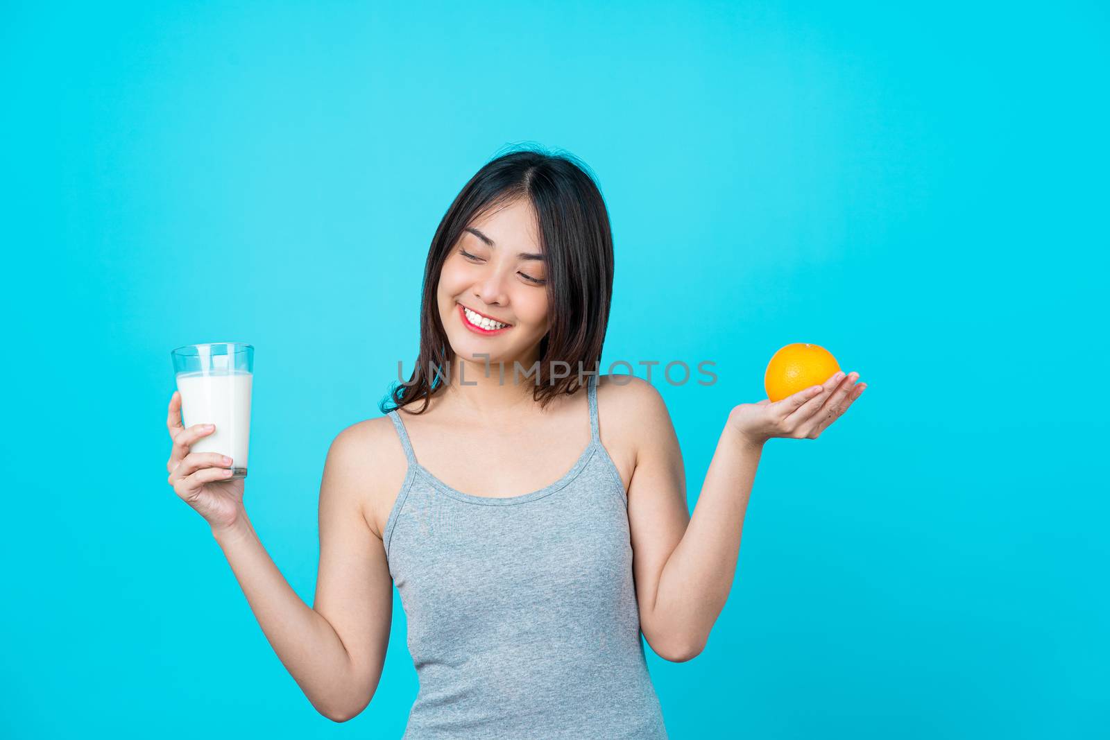 Attractive Asian young woman holding a glass of milk and a orange on isolated blue color background, weight loss and avoid junk food for dieting and Healthy