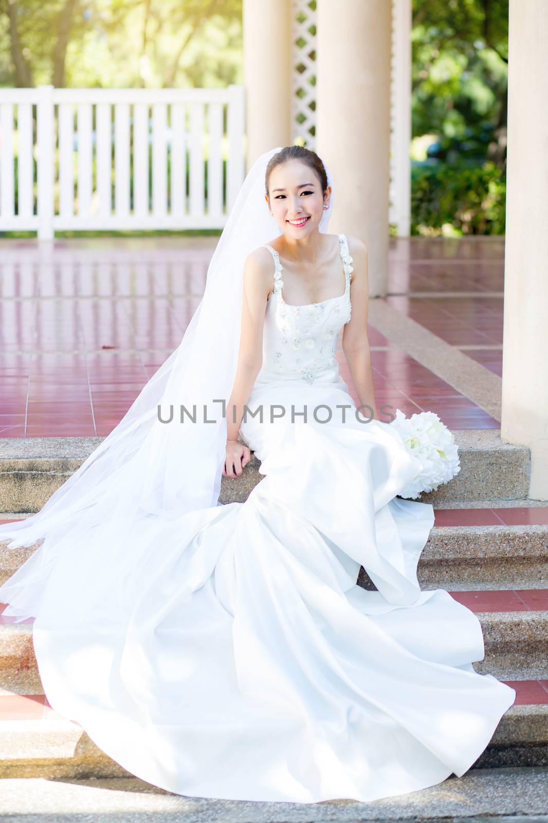 bride is sitting with flowers. Beautiful Young woman posing in park or garden in white bridal dress outdoors on a bright sunny day.