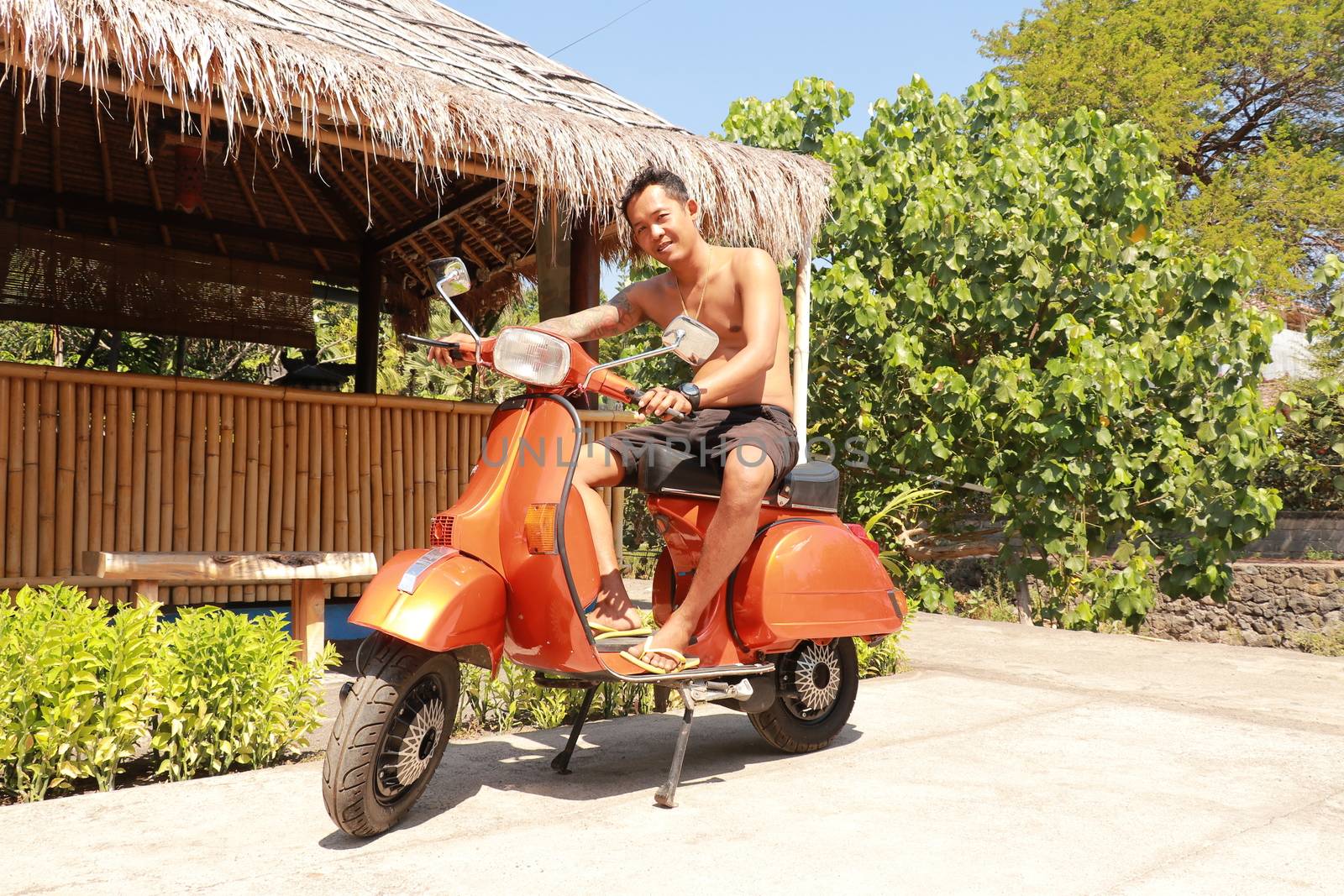 Bali, Indonesia, 1th of July, 2019. Youg Indonesia man is sitting on orange Classic Vespa. Orange Classic Vespa. A old orange Vespa Super motorscooter is parked on a sidewalk by Sanatana2008