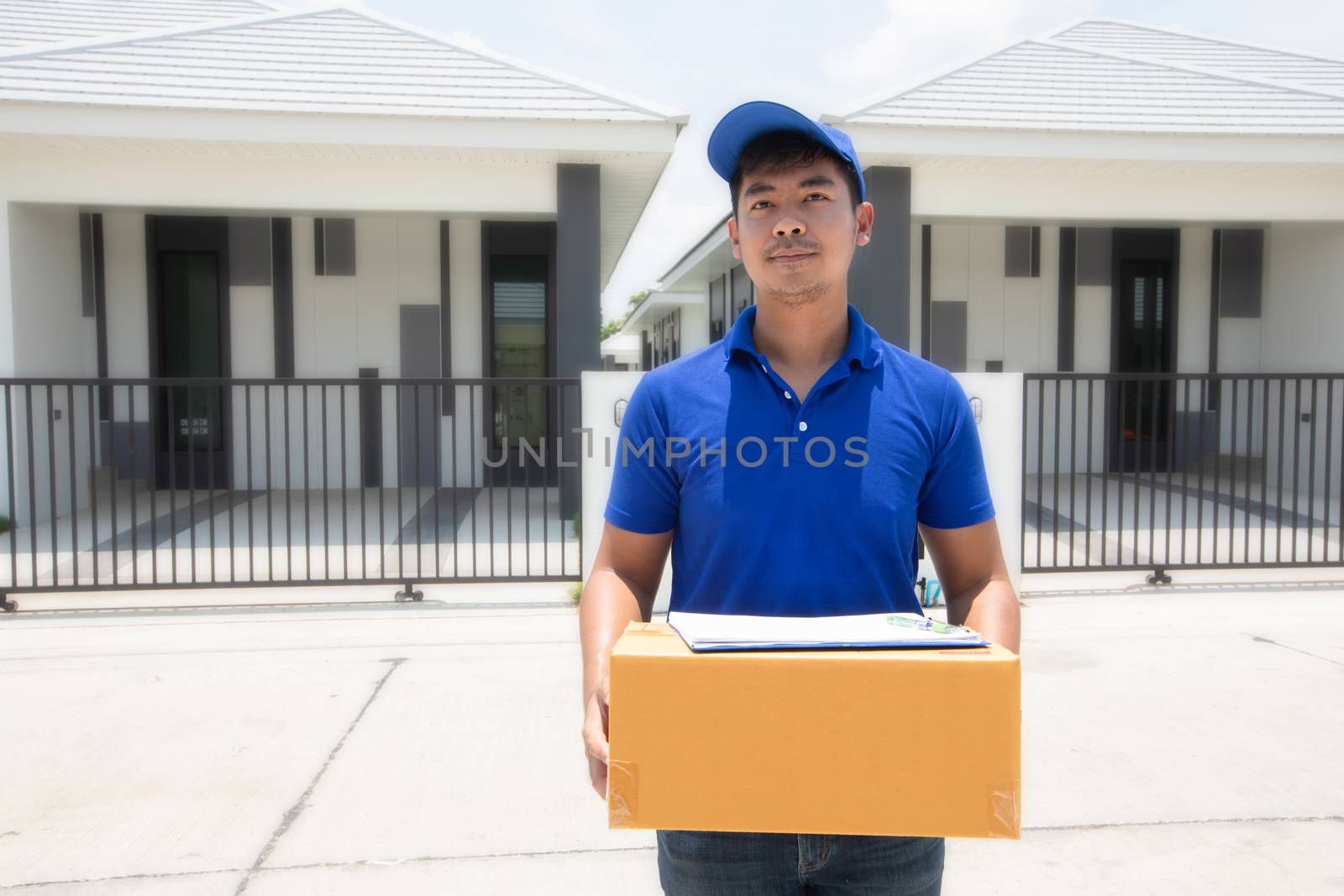 Asian delivery servicemen wearing a blue uniform with a bluecap and  handling cardboard boxes to give to the female customer in front of the house. Online shopping and Express delivery