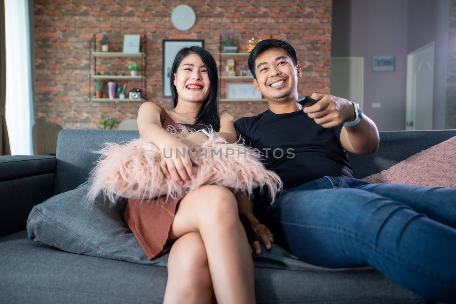 Asian couple is watching TV on the sofa at home on a holiday and smiling and laughing happy.