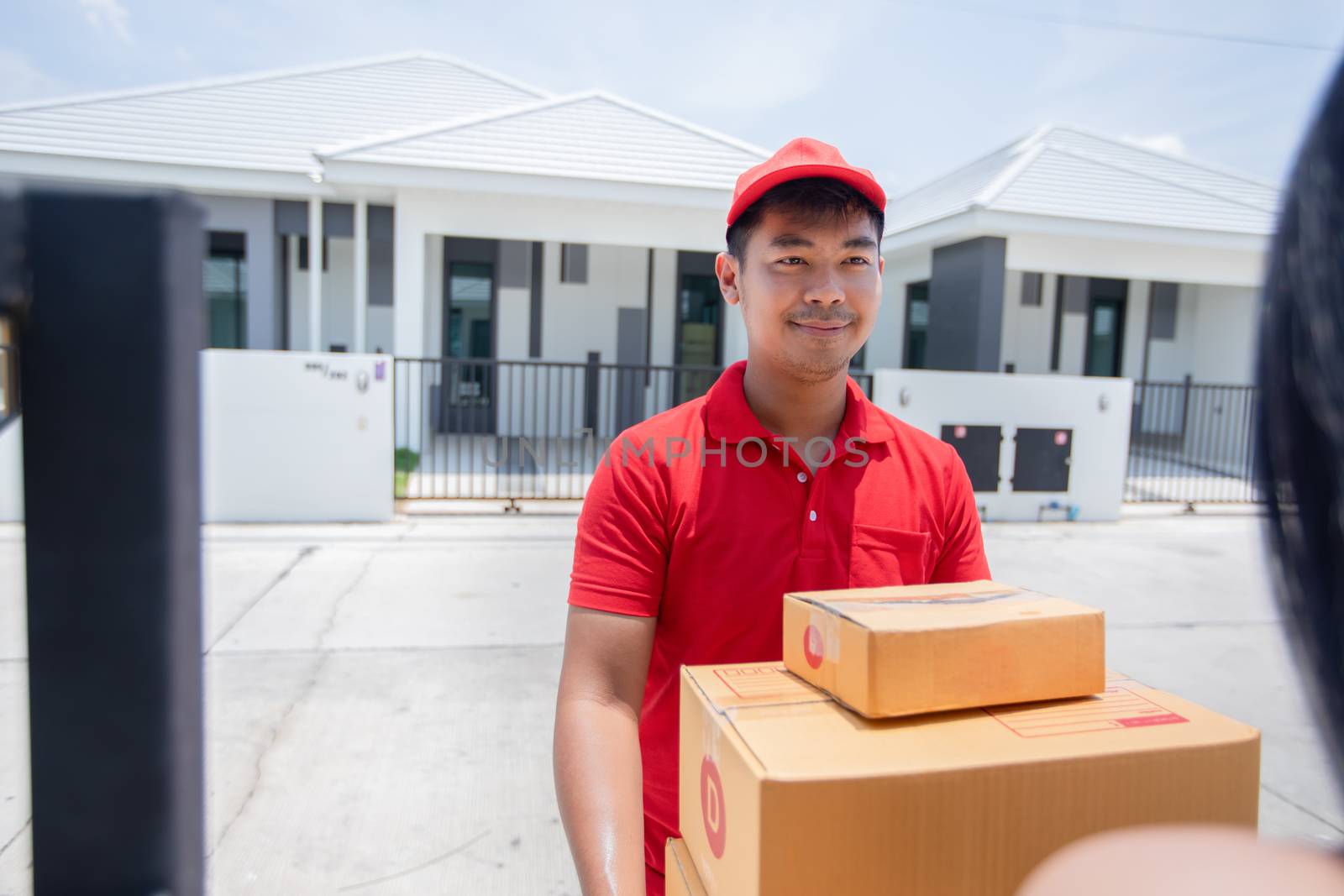 Asian delivery servicemen wearing a red uniform with a red cap and handling cardboard boxes to give to the female customer in front of the house. Online shopping and Express delivery