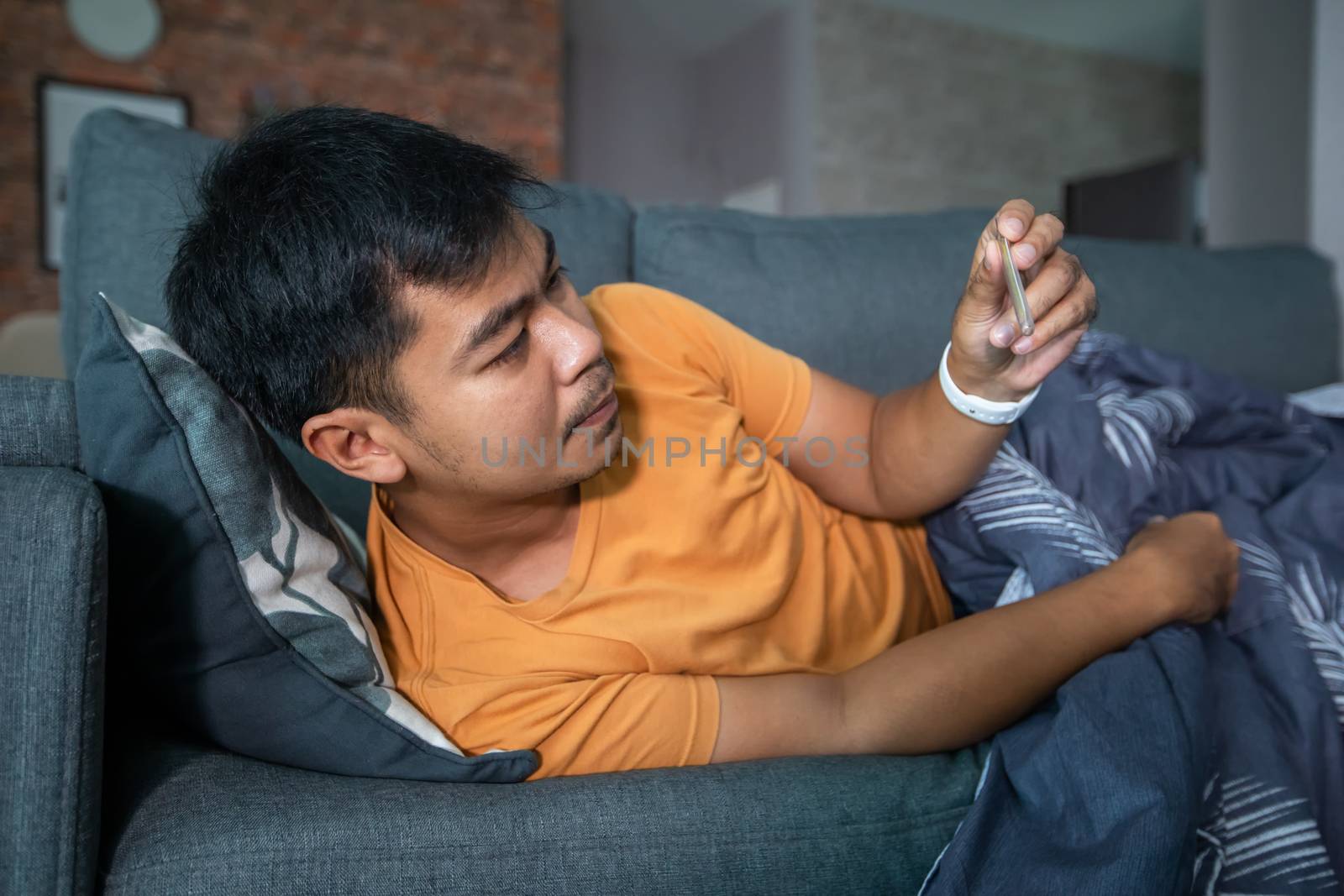 Asian man is ill and coughs on the sofa at home and he uses a thermometer to check the temperature during the coronavirus epidemic. by Tuiphotoengineer