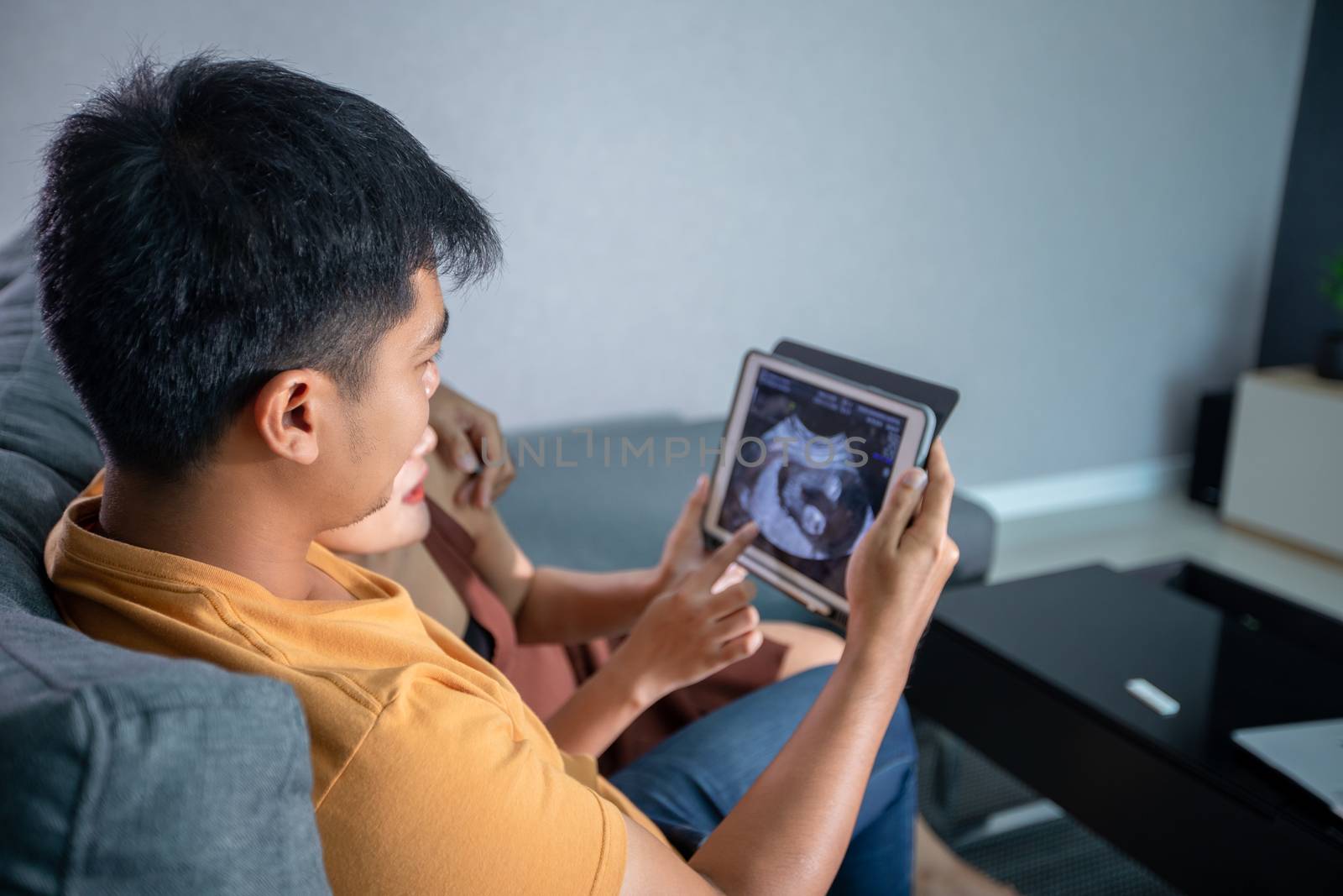 Asian Beautiful pregnant woman and husband looking ultrasound scan on a couch in the living room at home. Concept of Pregnancy health care.