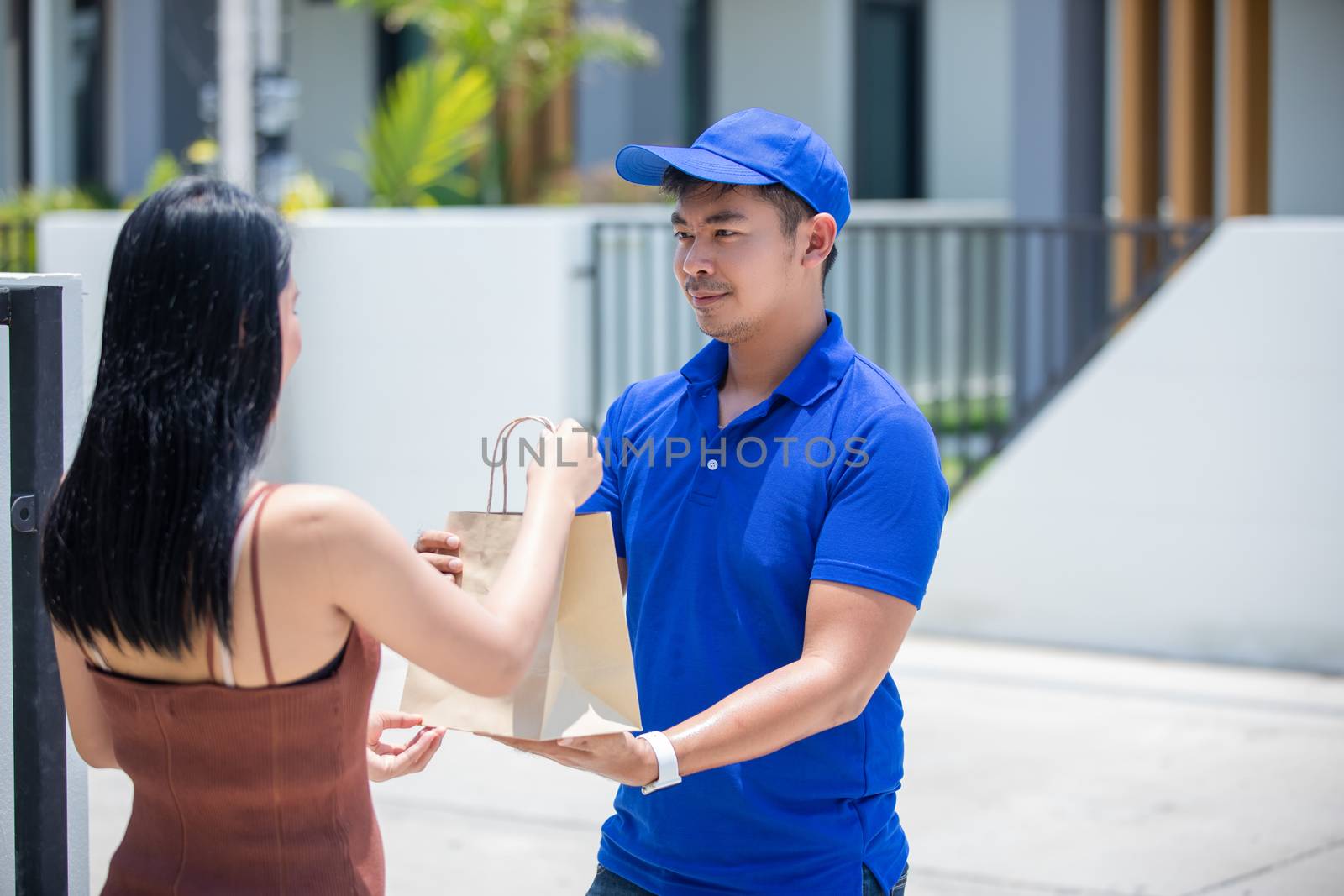 Asian delivery servicemen wearing a blue uniform with a bluecap and  handling cardboard boxes to give to the female customer in front of the house. Online shopping and Express delivery by Tuiphotoengineer