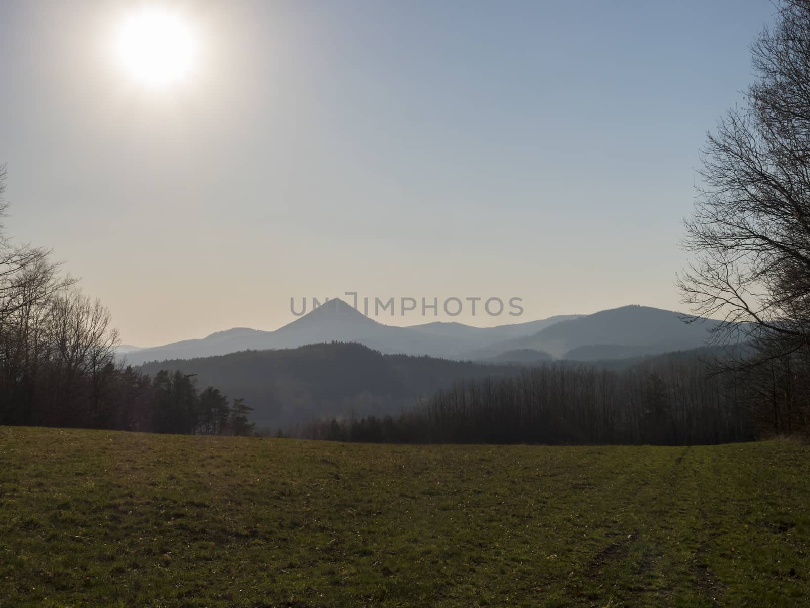 early spring landscape at Lusatian mountains, with view point hill klic, grass meadow, bare trees, deciduous and spruce tree forest, clear blue sky background, golden hour light, horizontal, copy space.