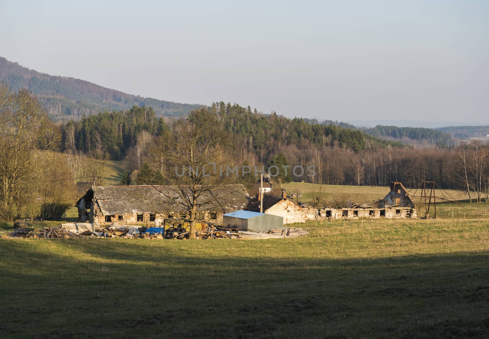 Old ruin of abandoned farm house at Lusatian mountains, early spring landscape with grass meadow, bare trees, forest, hills, clear blue sky background, golden hour light, horizontal, copy space by Henkeova