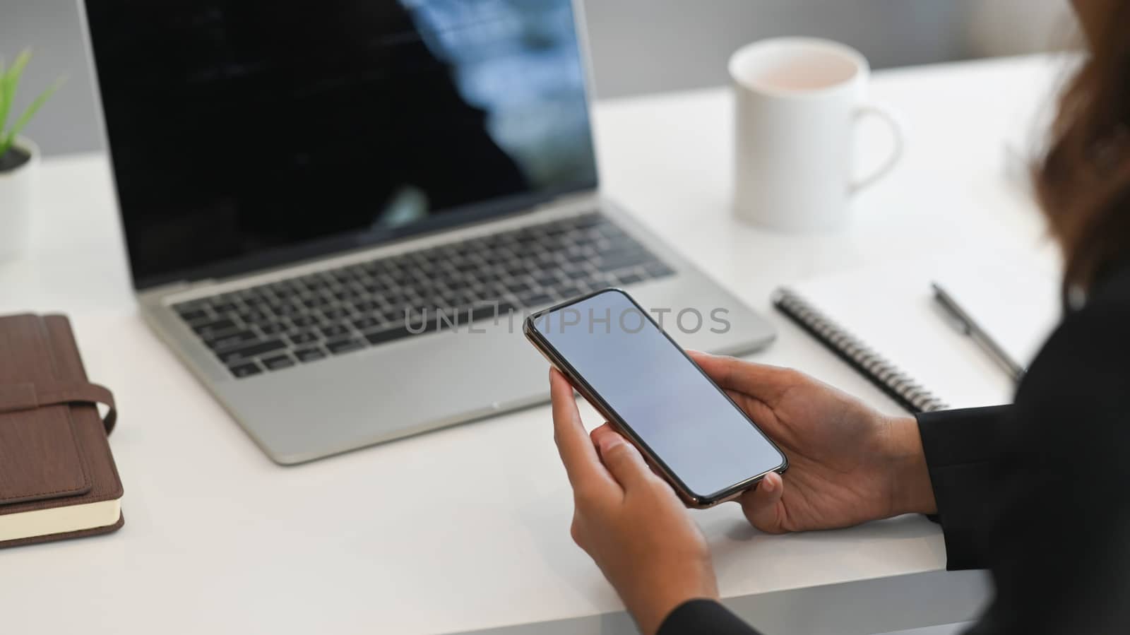 Cropped shot of businesswoman while holding blank screen mobile in hand on the working desk. Black blank screen laptop, notebook, coffee cup and document on the working desk.