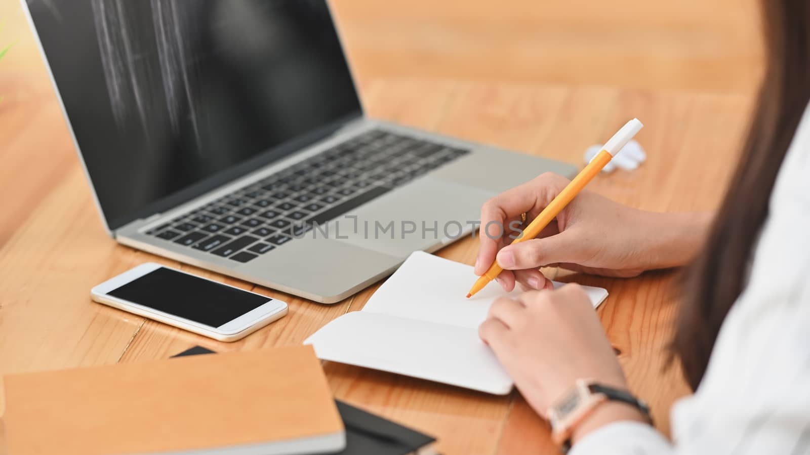 Cropped shot of young business woman while take notes in front of the black screen laptop on the  wooden working desk at the work place. 