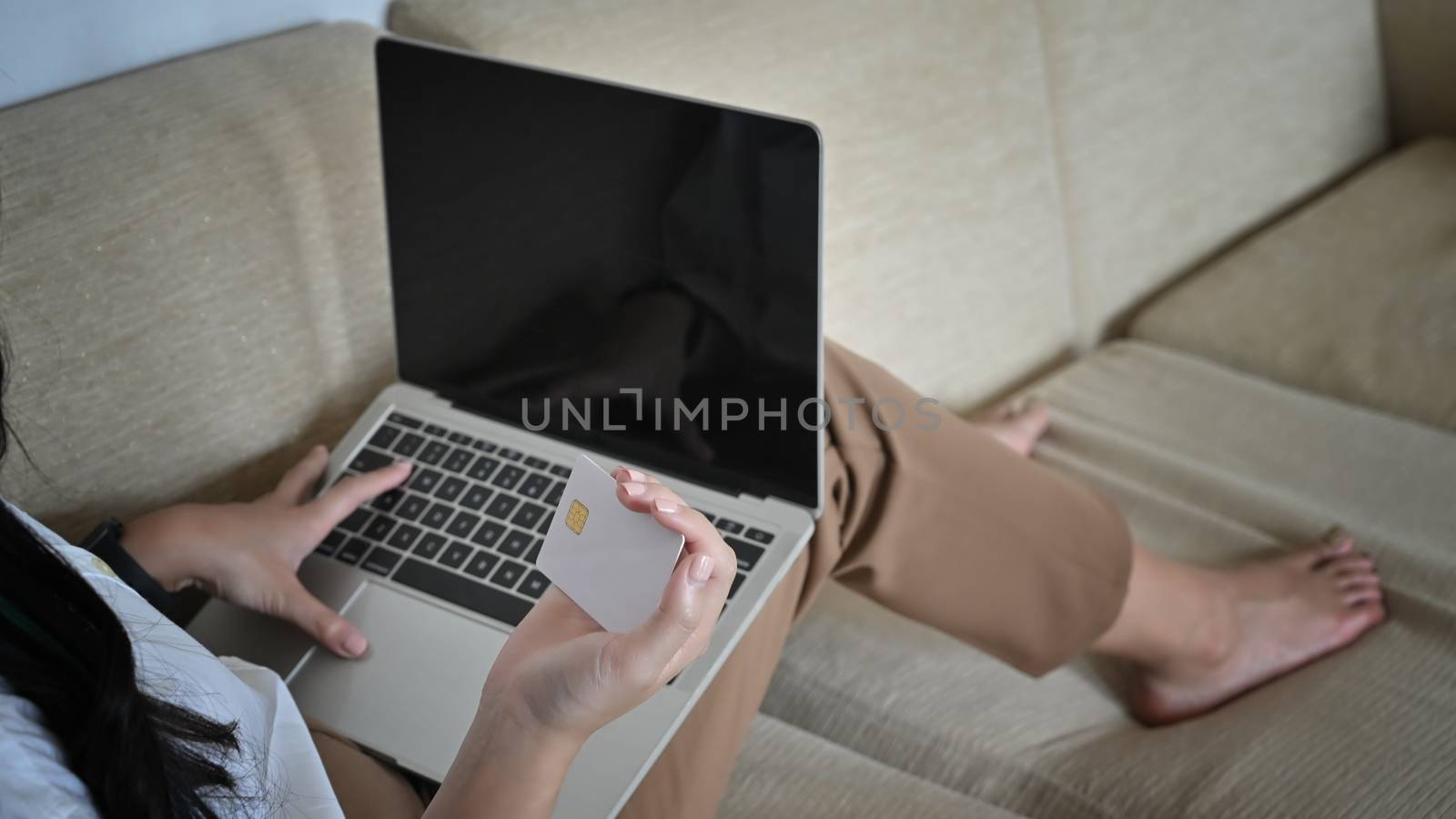 Close-up young relaxing beautiful woman sitting on the sofa while typing on the keyboard and  looking at her personal card. Fill the information from card.