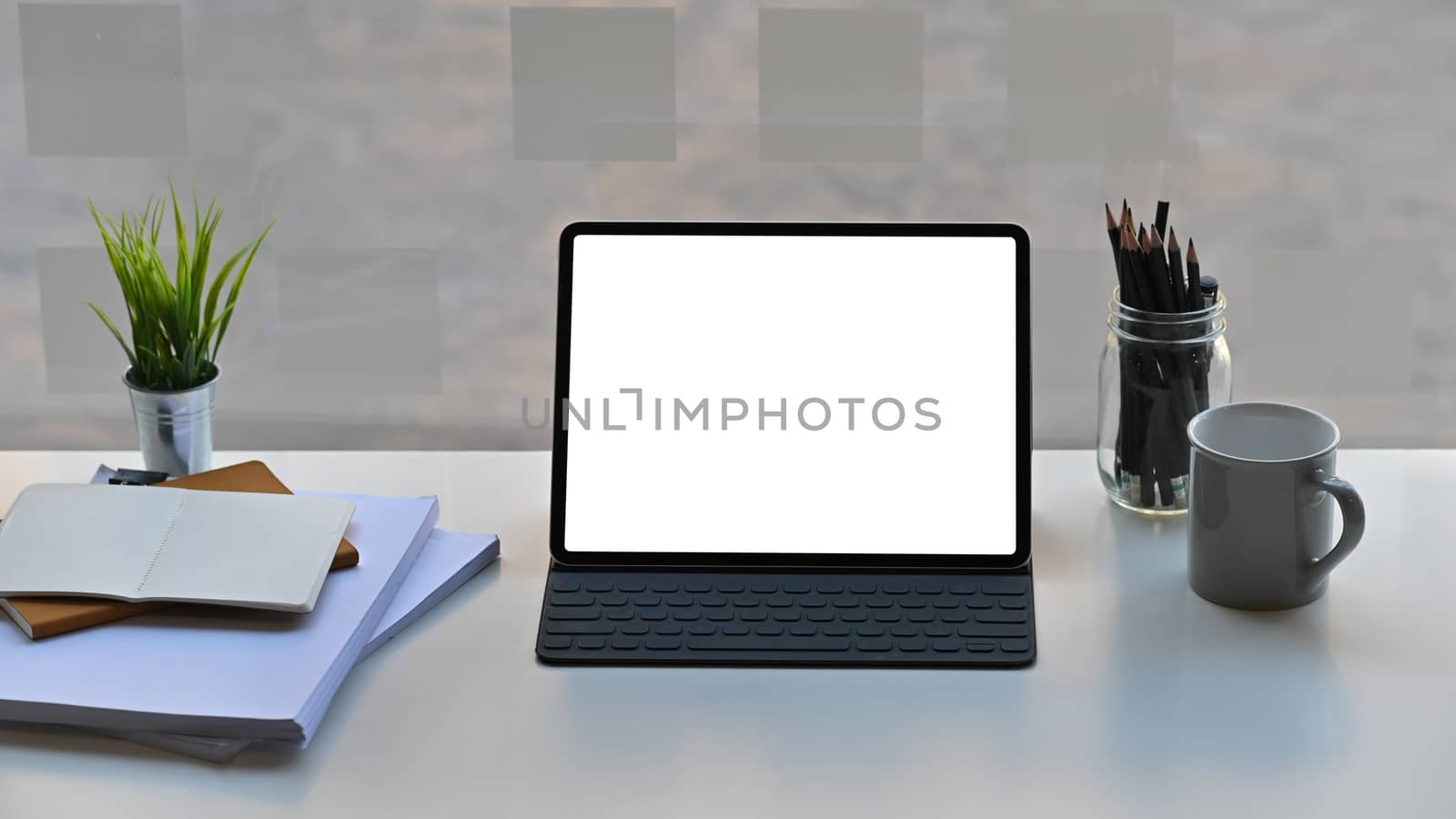 Photo of white blank screen computer tablet in a leather-case and keyboard is setting on a modern working table over comfortable office background.