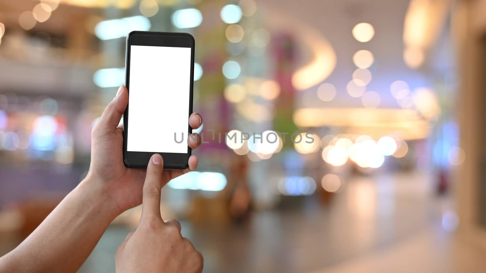 Close-up image of hand while holding crop black smartphone with white blank screen display and touching on home button over the shopping center blurred background.