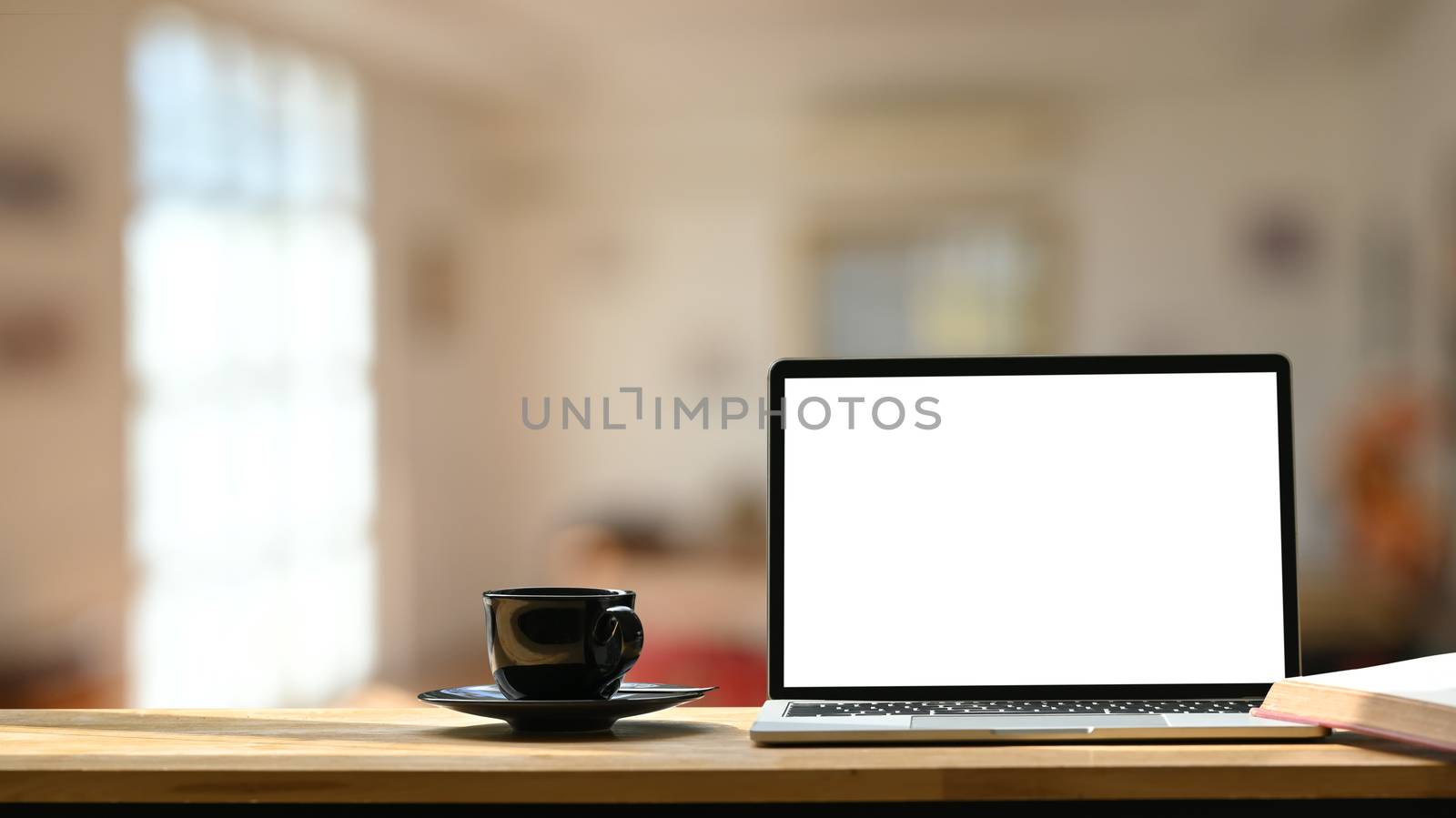 Photo of white blank screen laptop, black coffee cup and coasters on the wooden working desk over blurred modern cafe background.