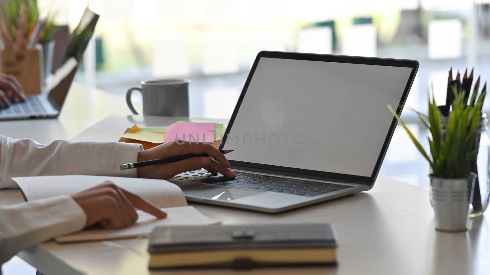 Close-up image of businesswoman typing on white blank screen laptop and copying information from document to laptop at the modern working table.