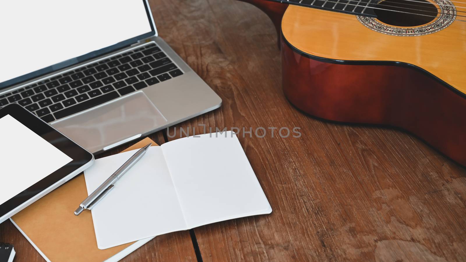 Work table of songwriter. Cropped shot of note, pencil, acoustic guitar, white blank screen tablet and laptop all of these is putting on the wooden desk.