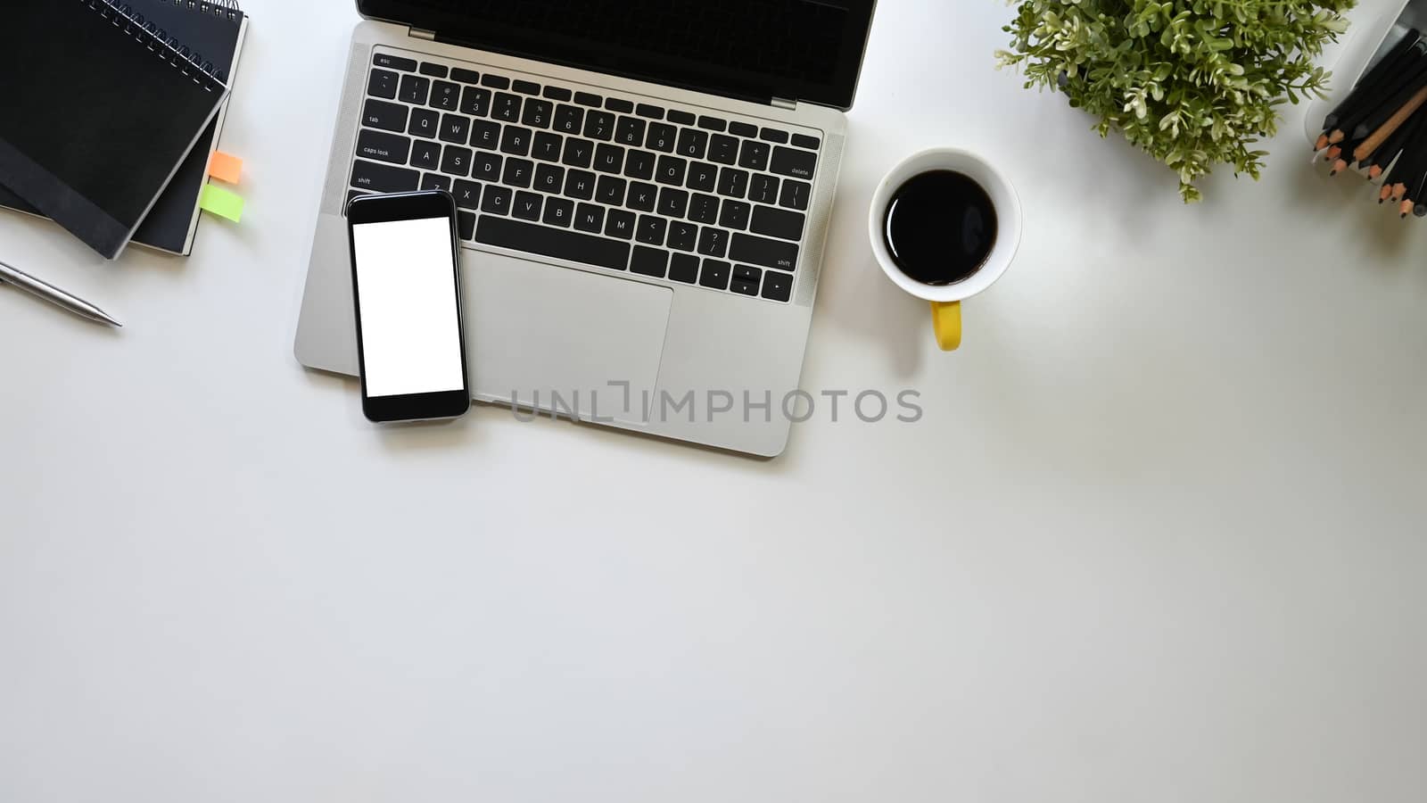 Top view office desk including a coffee cup, laptop, white blank screen smartphone, potted plant, pen and notebook putting on it. Office equipment with flat laid.