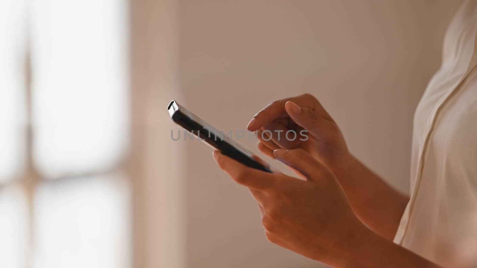 Side shot of beautiful businesswoman in white shirt surfing the internet by using smartphone in her hand with modern minimal living room as background.