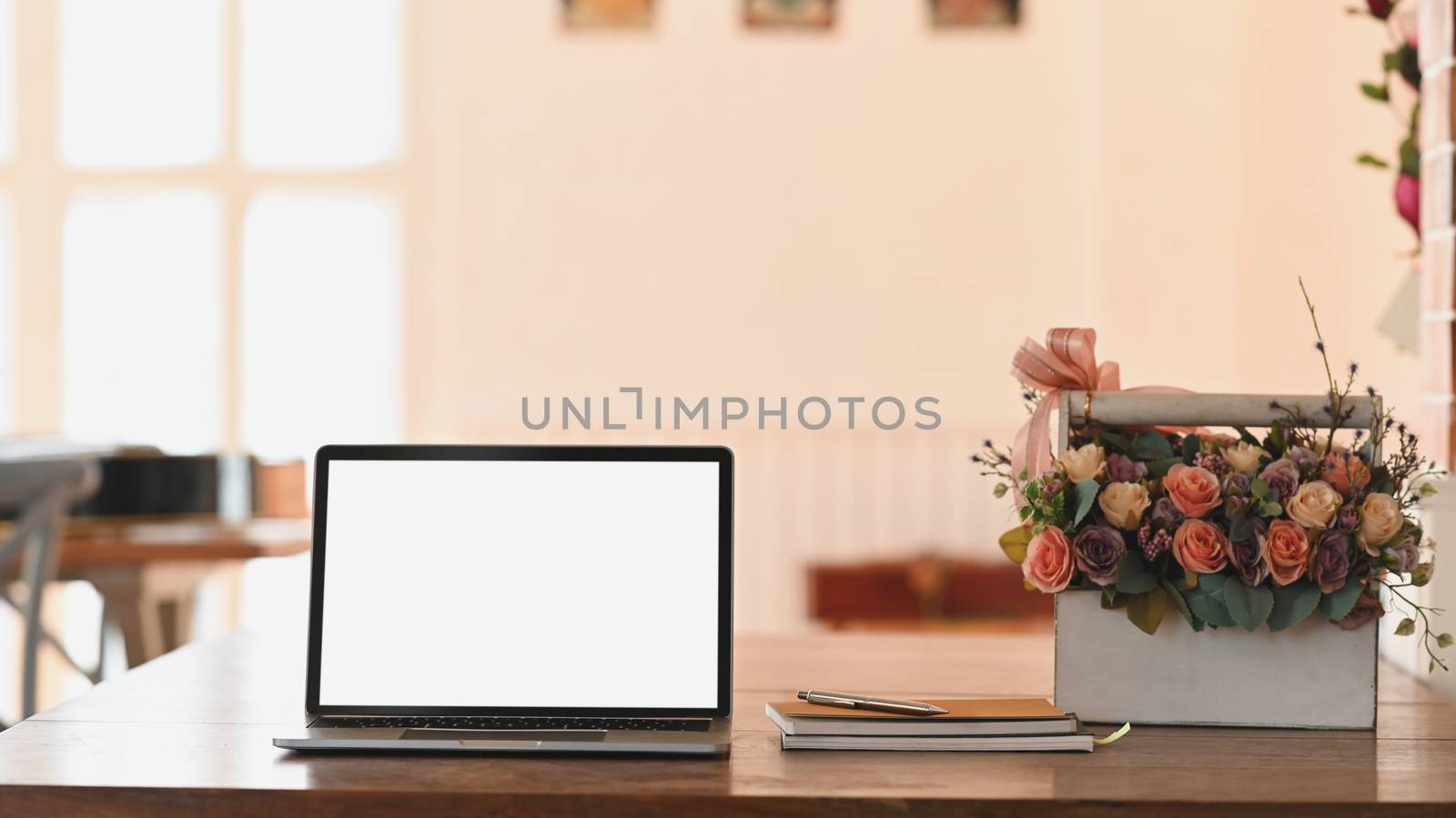 Blank screen laptop, notebook and roses bouquet in white plastic basket all of these putting on the wooden table with modern living room as background.