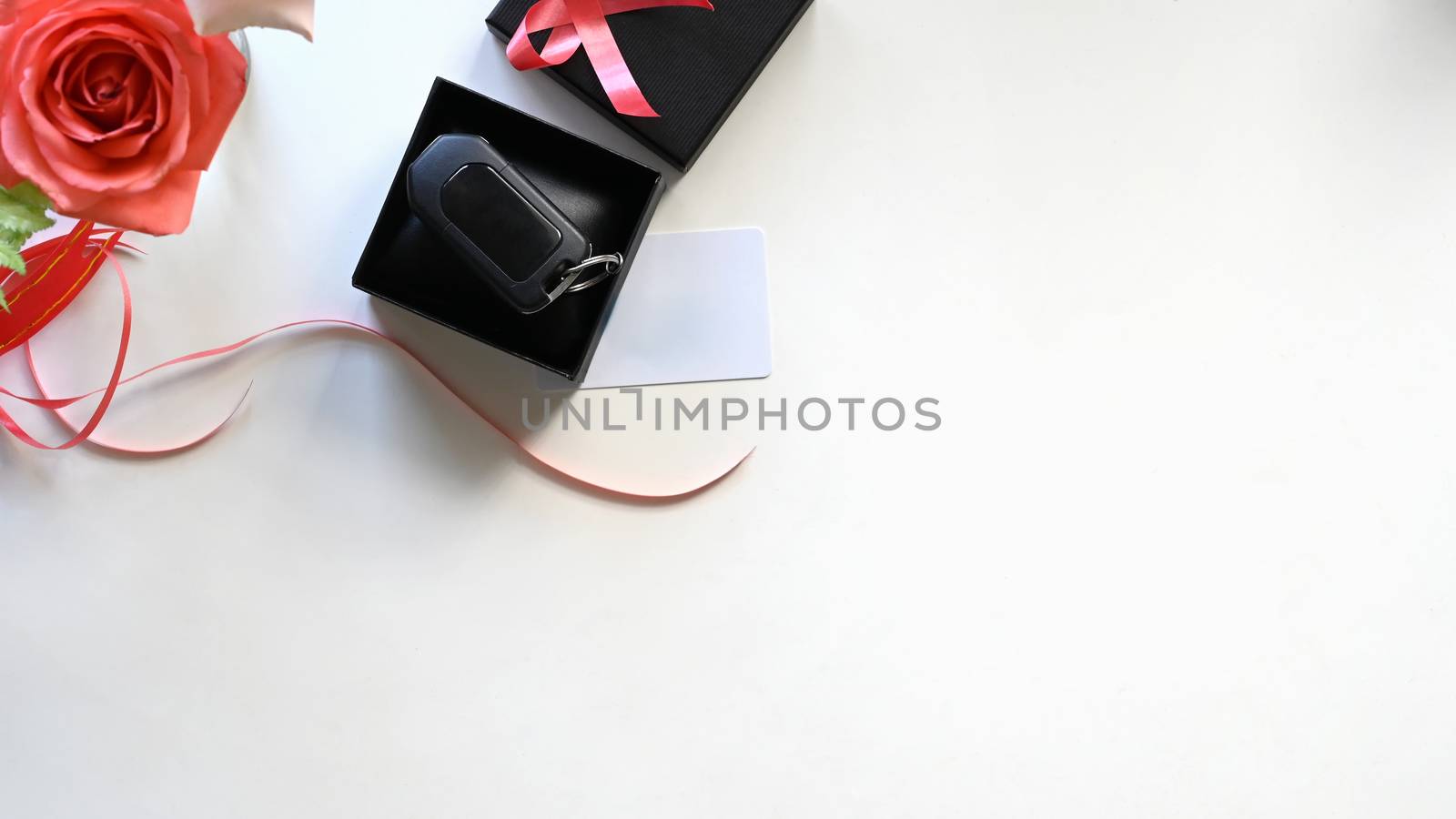 Top view Digital car key putting inside the black gift box with red ribbon,bouquet of roses and wish card on the white desk as background. Surprising Valentine's Day gift concept.