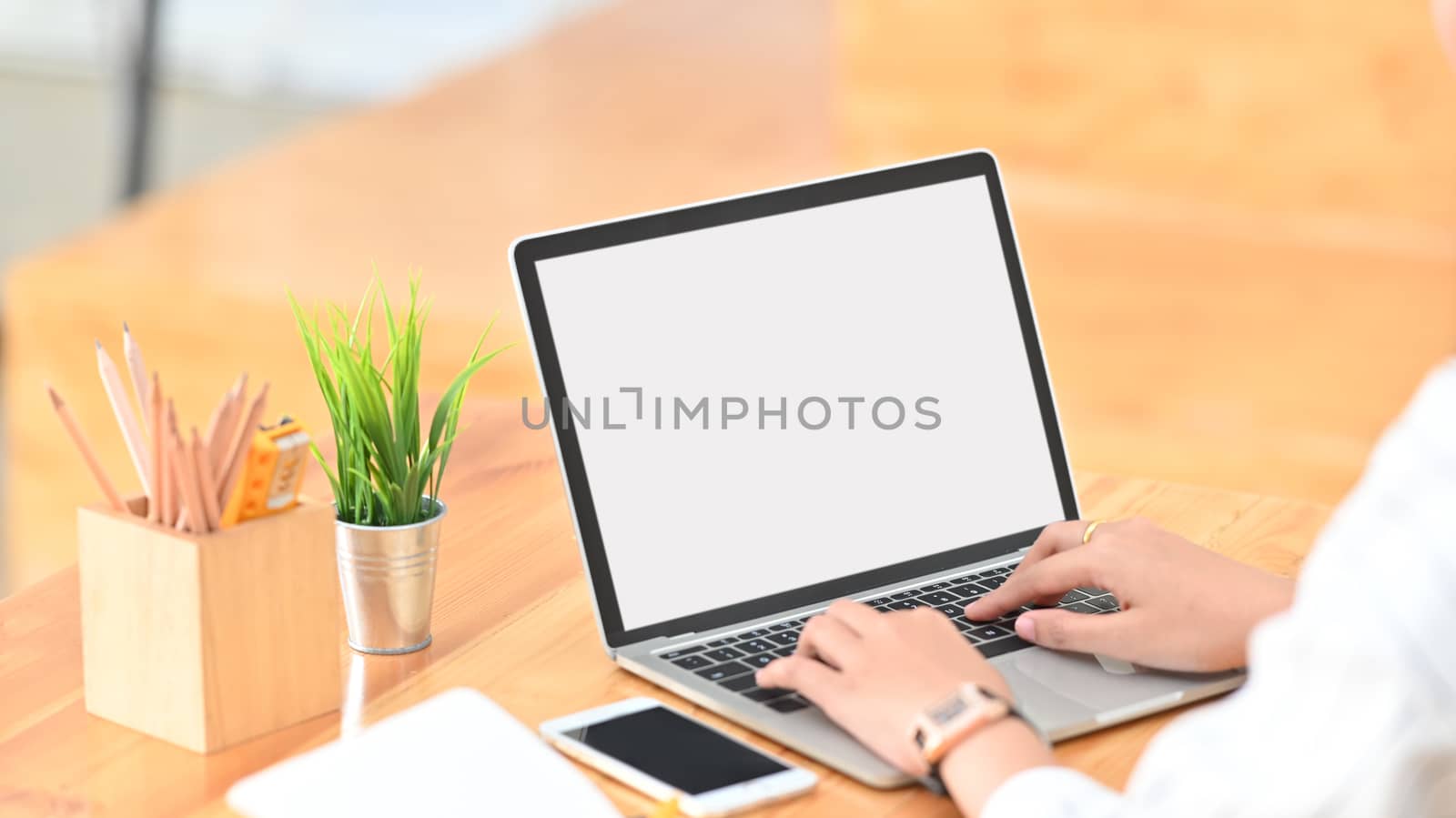 Cropped shot of young creative woman surfing the internet by using white blank screen laptop while sitting at the modern wooden desk.