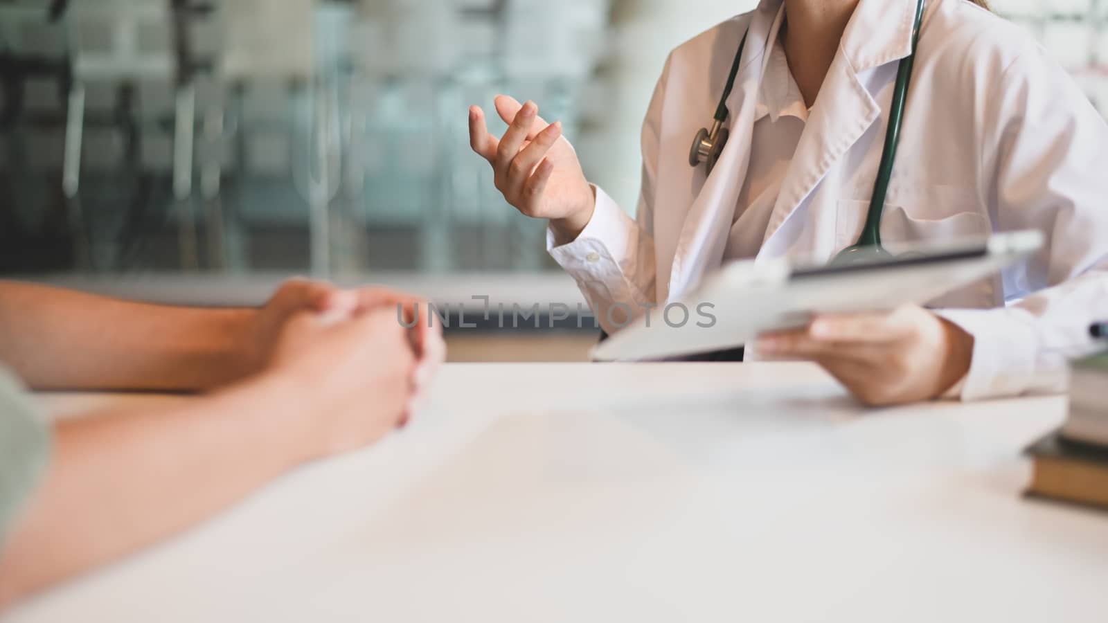 Cropped shot of woman working doctor while giving an illness information to her patient at the white working desk with the modern hospital as background.