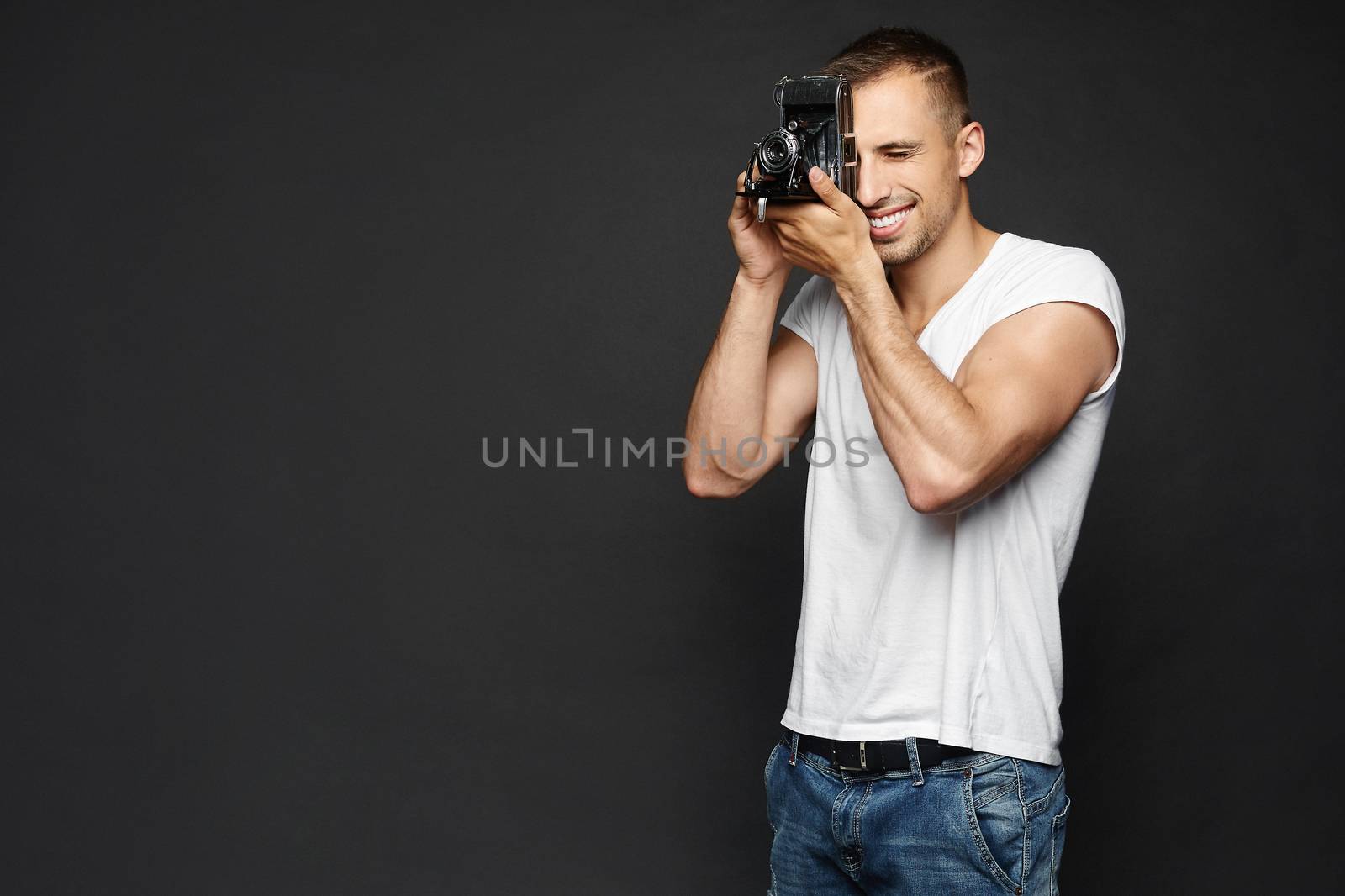 Young smiling handsome man holding a vintage photo camera and making a photoshoot, isolated at the dark background.