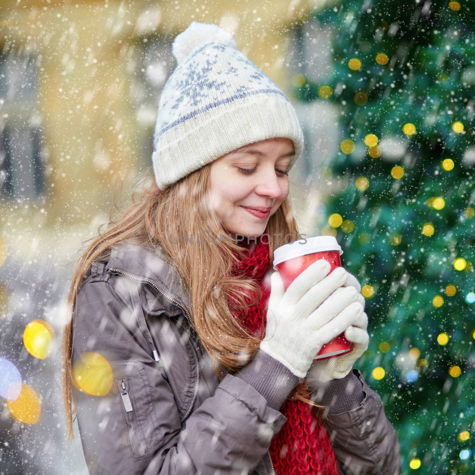Girl drinking take away coffee, hot chocolate or eggnog near decorated Christmas tree during snowfall