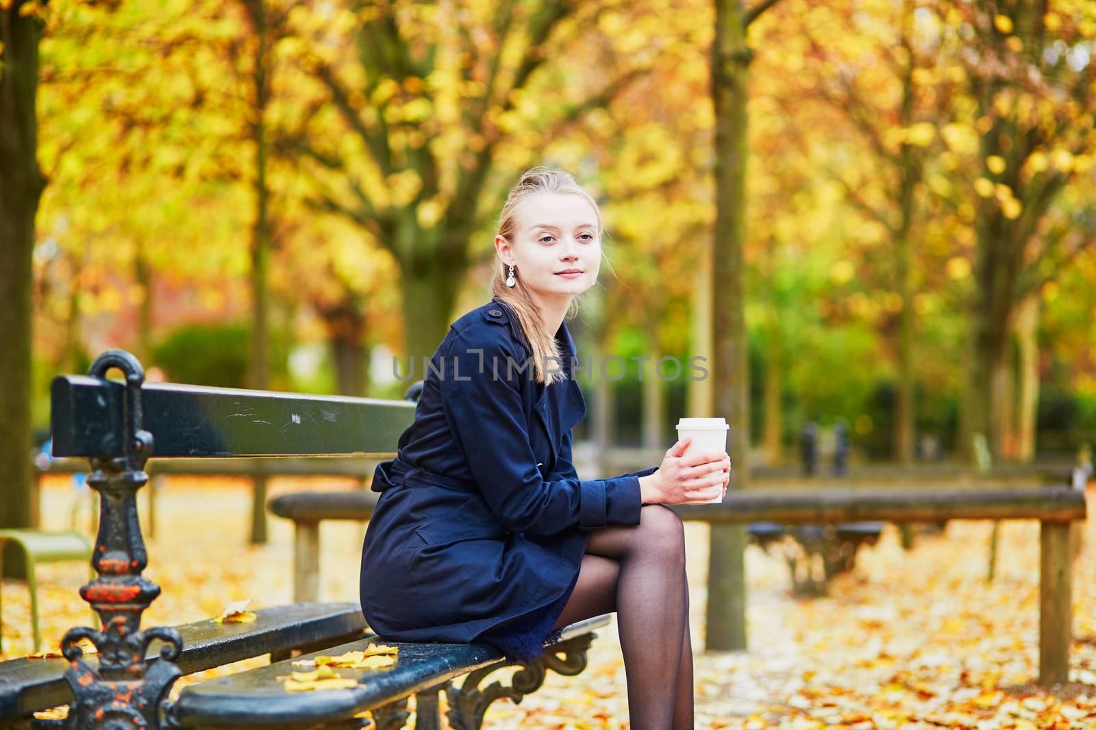 Beautiful young woman in the Luxembourg garden of Paris on a fall day, drinking coffee to go and reading book