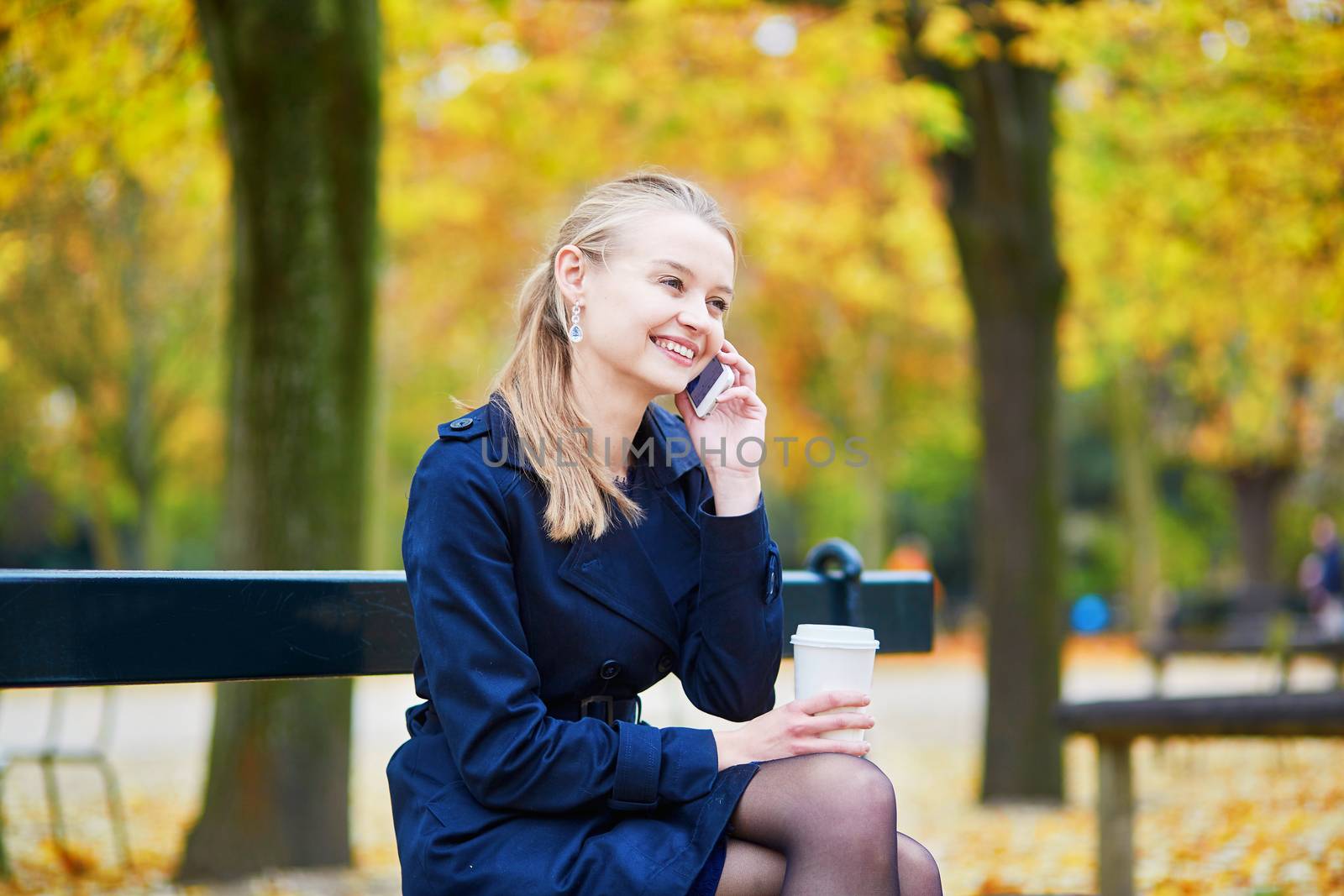 Beautiful young woman drinking coffee and speaking on the phone in the Luxembourg garden of Paris on a fall day