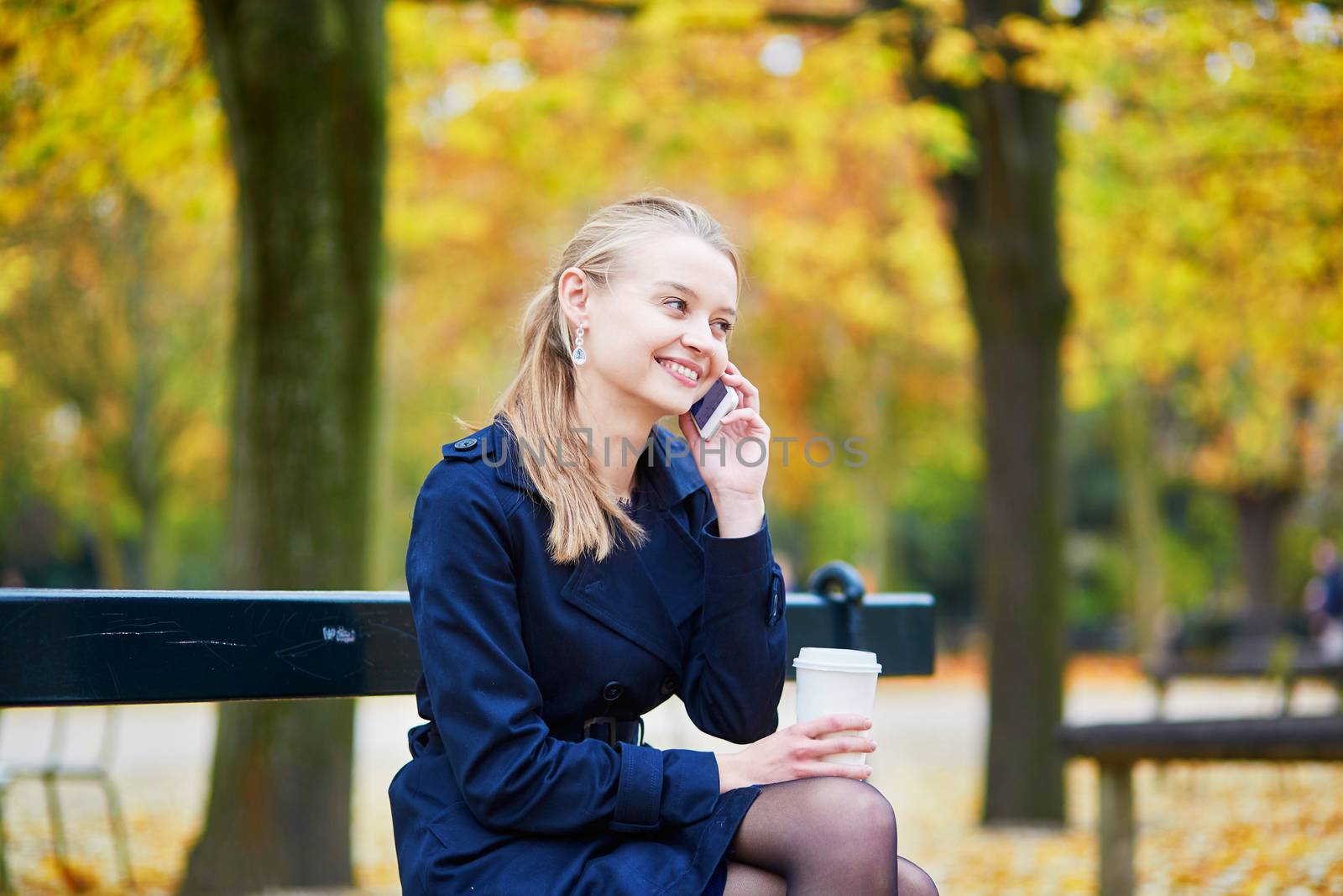 Beautiful young woman in the Luxembourg garden of Paris on a fall day, drinking coffee to go and using mobile phone