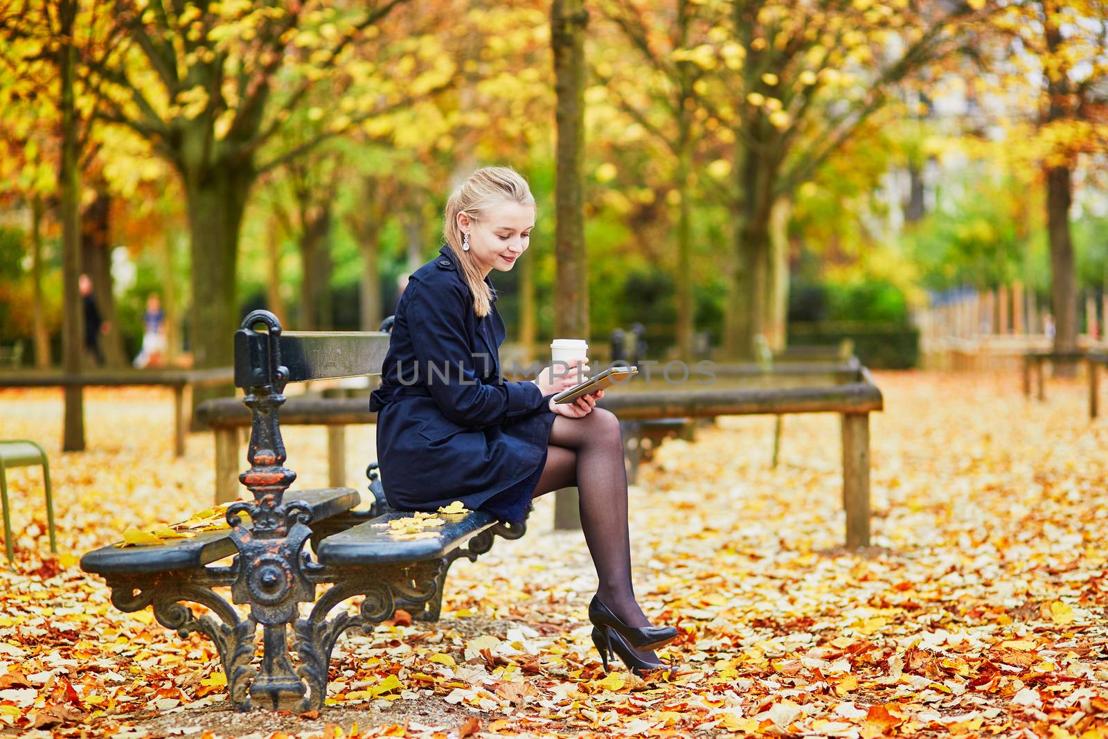 Beautiful young woman in the Luxembourg garden of Paris on a fall day, drinking coffee to go and reading book