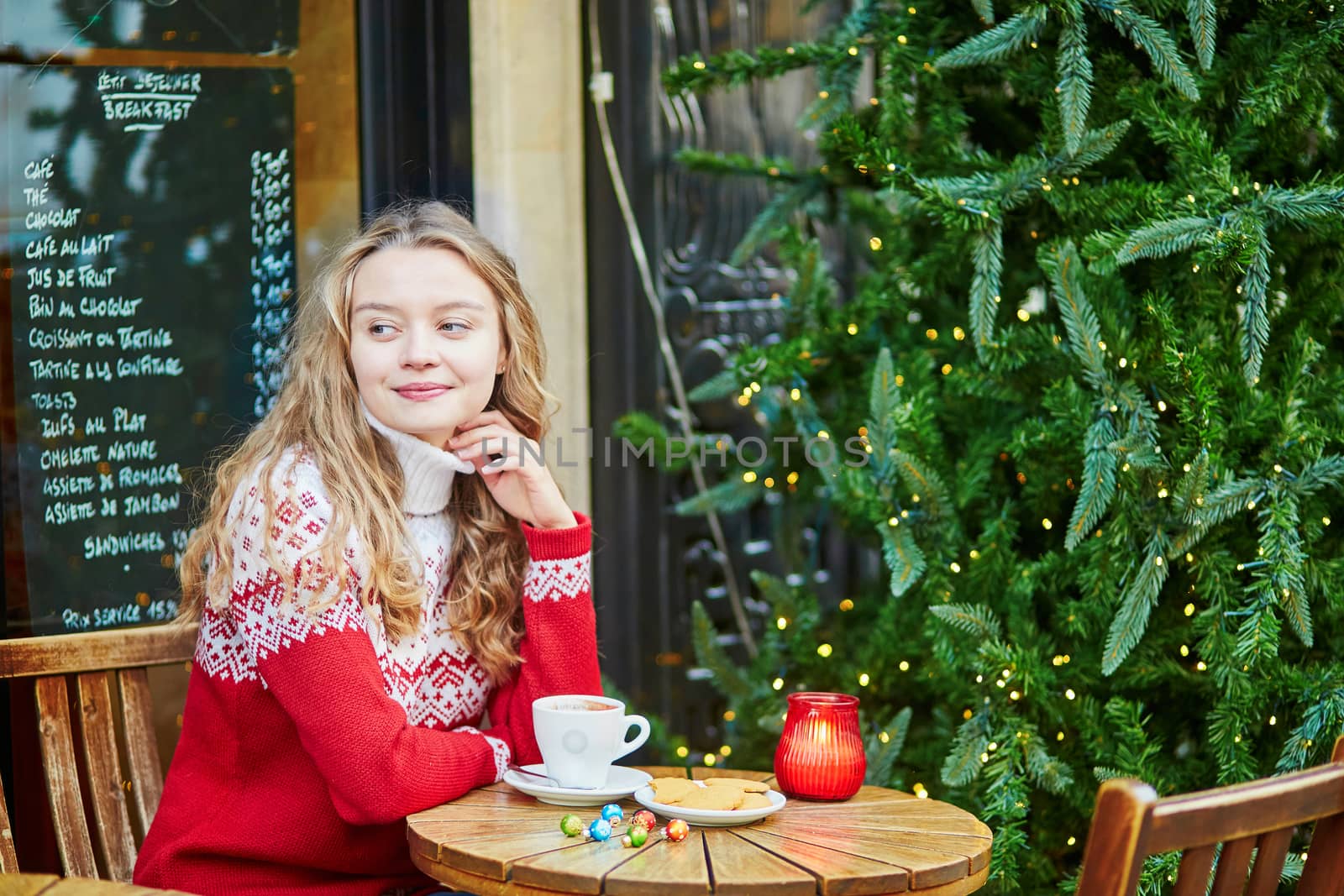 Cheerful young woman drinking hot chocolate in a cozy outdoor Parisian cafe decorated for Christmas