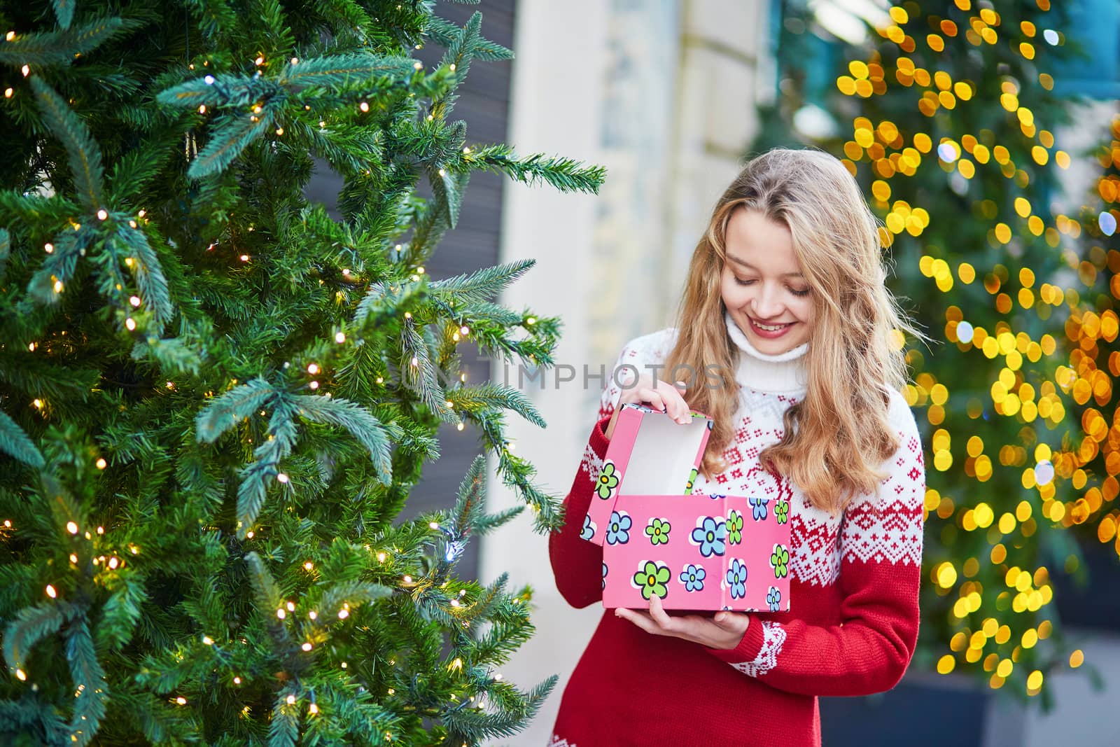 Cheerful young woman on a street of Paris decorated for Christmas, unpacking Christmas present