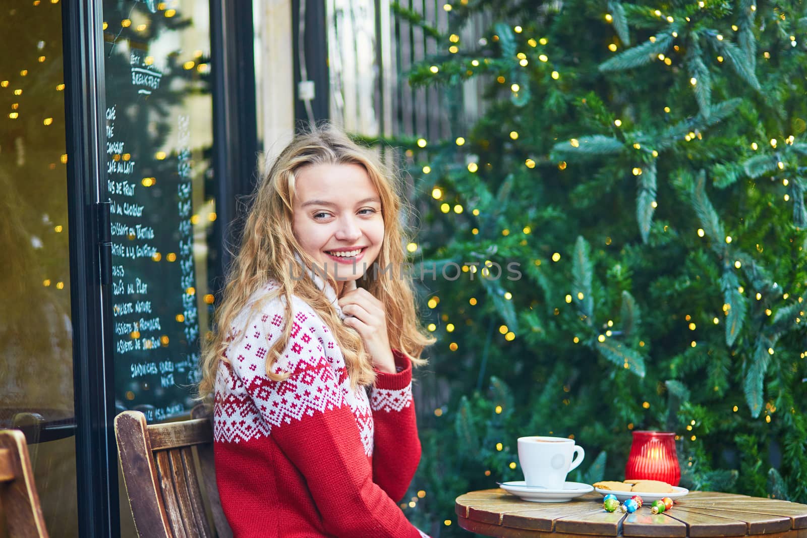 Cheerful young woman drinking hot chocolate in a cozy outdoor Parisian cafe decorated for Christmas