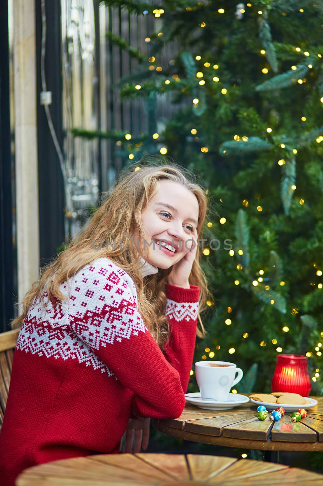 Cheerful young woman drinking hot chocolate in a cozy outdoor Parisian cafe decorated for Christmas
