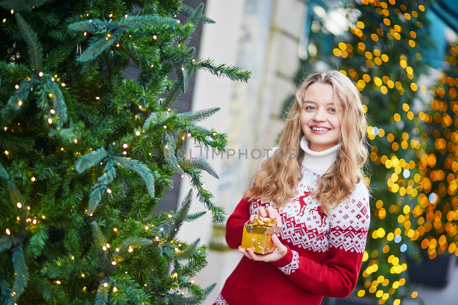 Cheerful young woman on a street of Paris decorated for Christmas, unpacking Christmas present