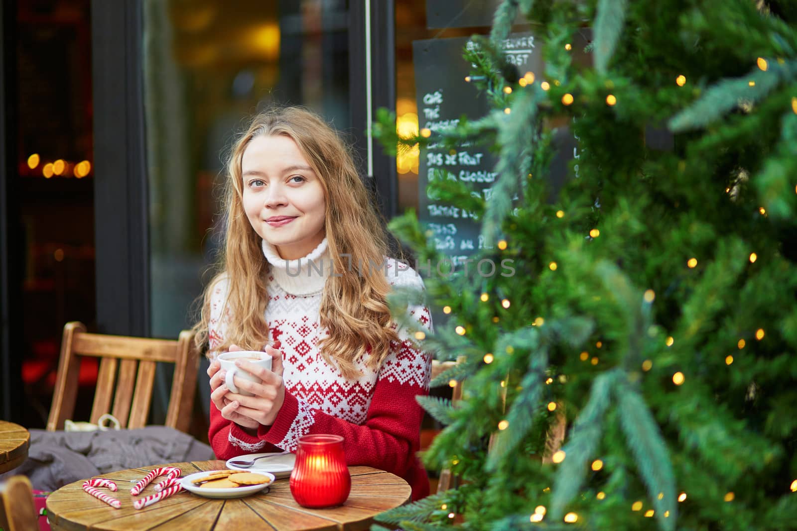 Cheerful young woman drinking hot chocolate in a cozy outdoor Parisian cafe decorated for Christmas