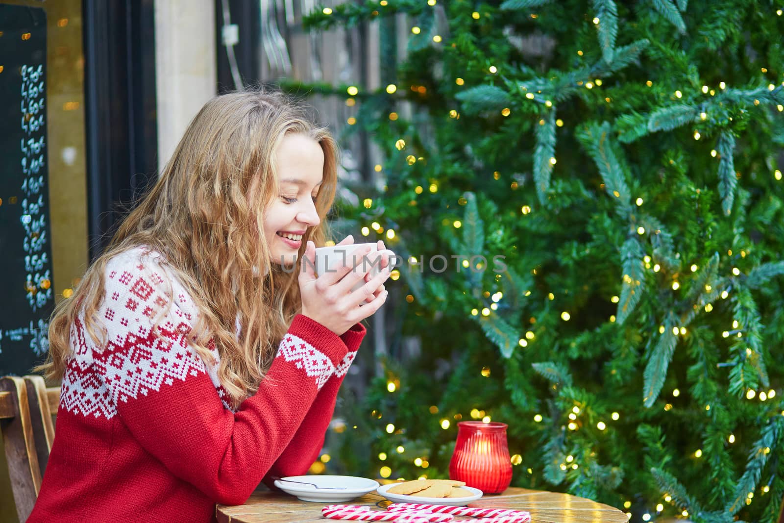 Cheerful young woman drinking hot chocolate in a cozy outdoor Parisian cafe decorated for Christmas