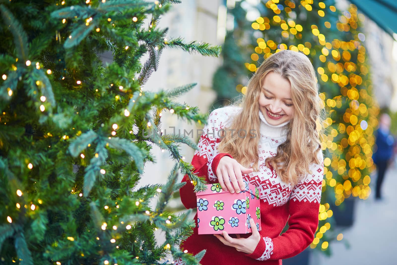 Cheerful young woman on a street of Paris decorated for Christmas, unpacking Christmas present