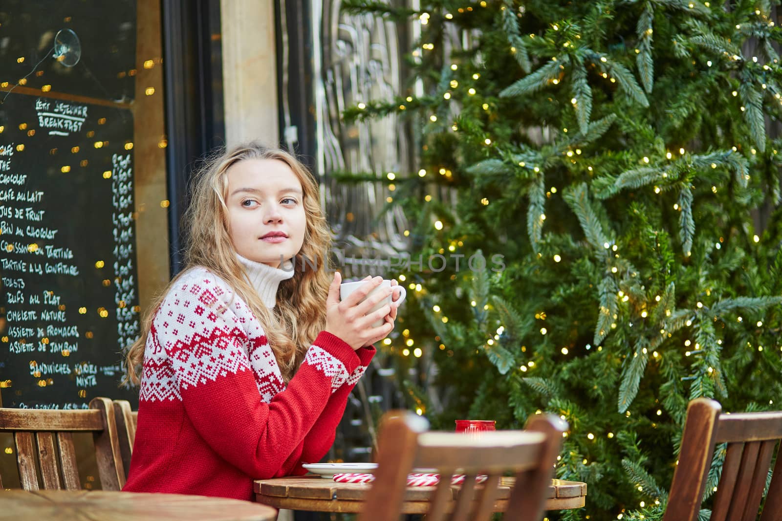 Cheerful young woman drinking hot chocolate in a cozy outdoor Parisian cafe decorated for Christmas
