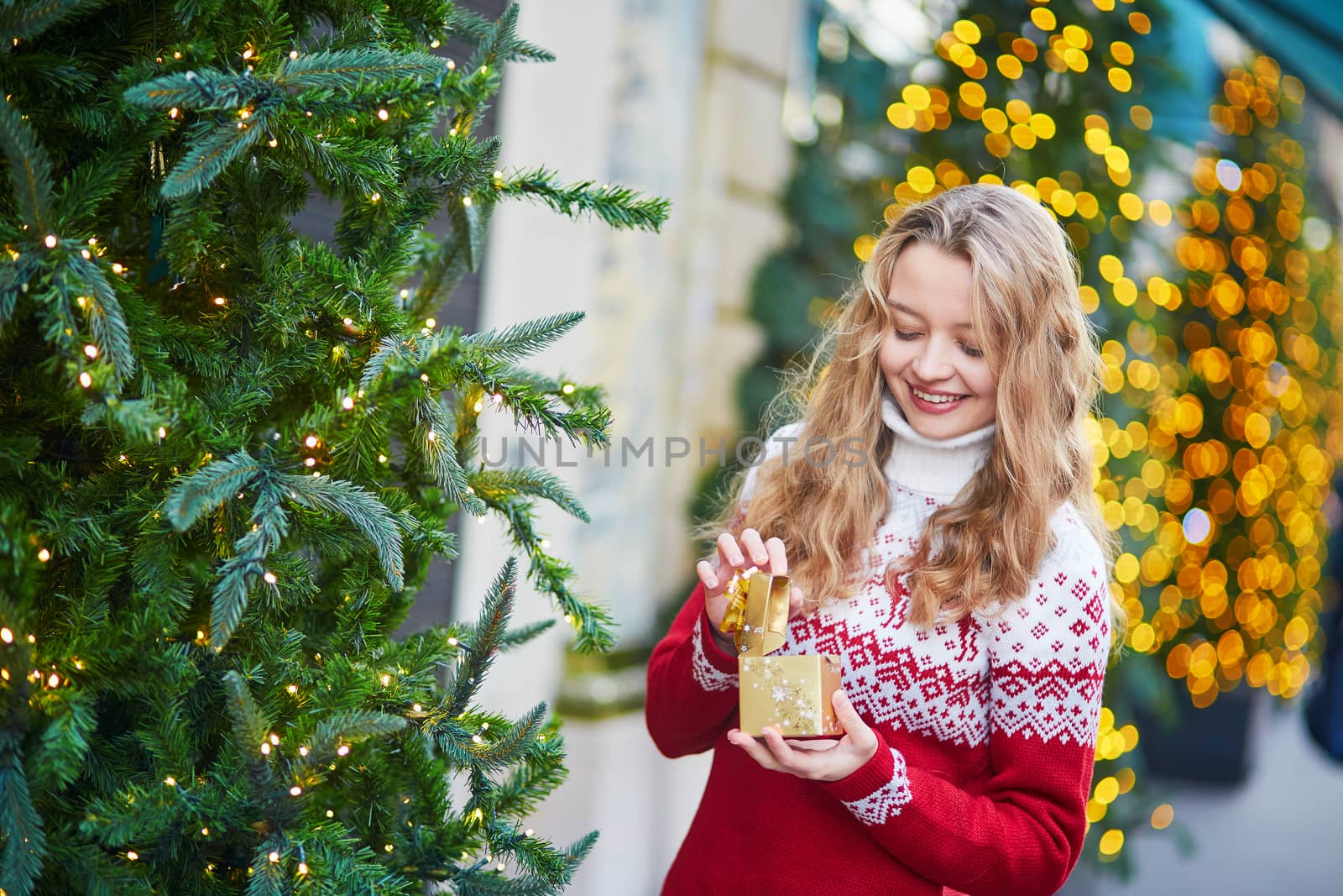 Cheerful young woman on a street of Paris decorated for Christmas, unpacking Christmas present