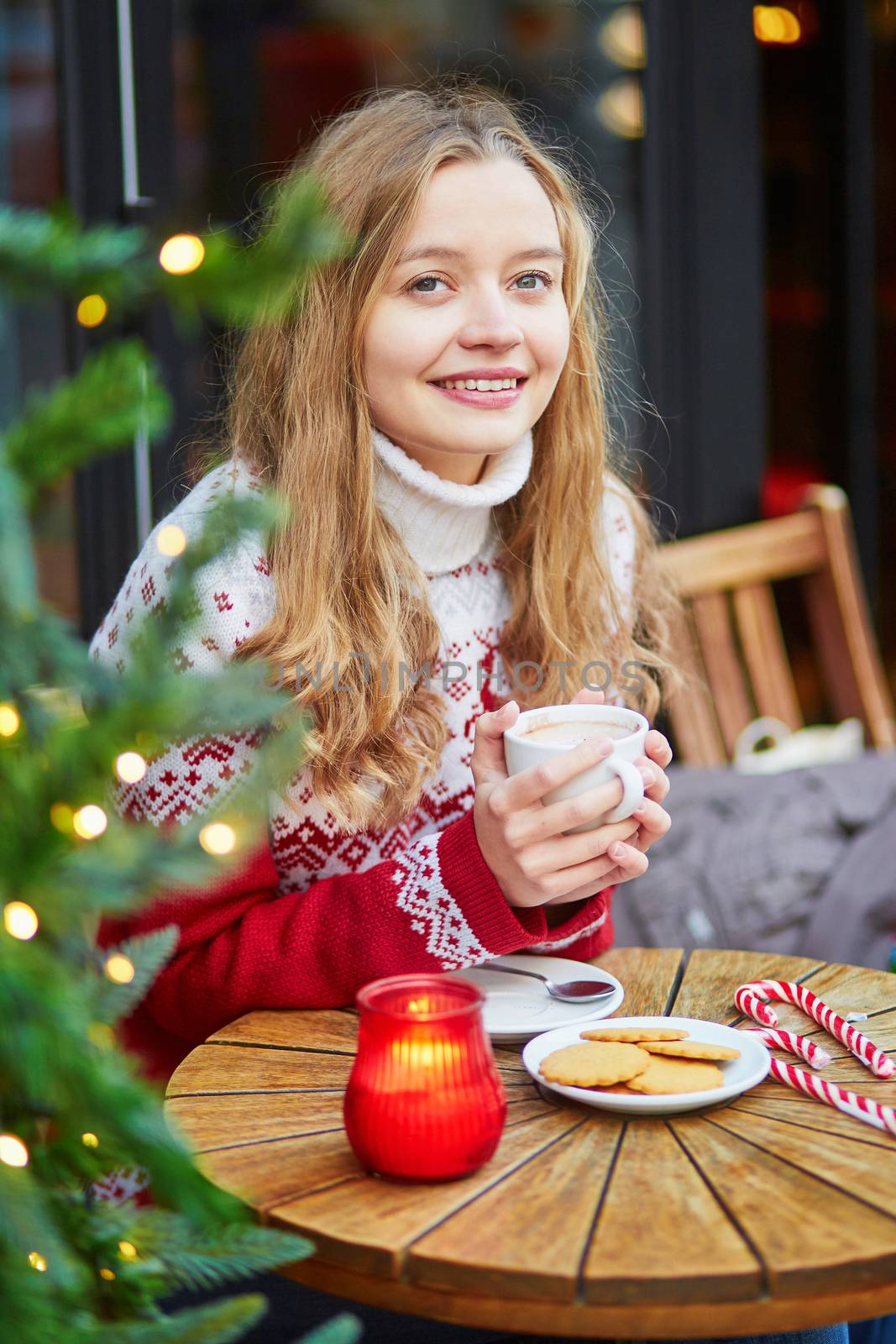 Cheerful young woman drinking hot chocolate in a cozy outdoor Parisian cafe decorated for Christmas
