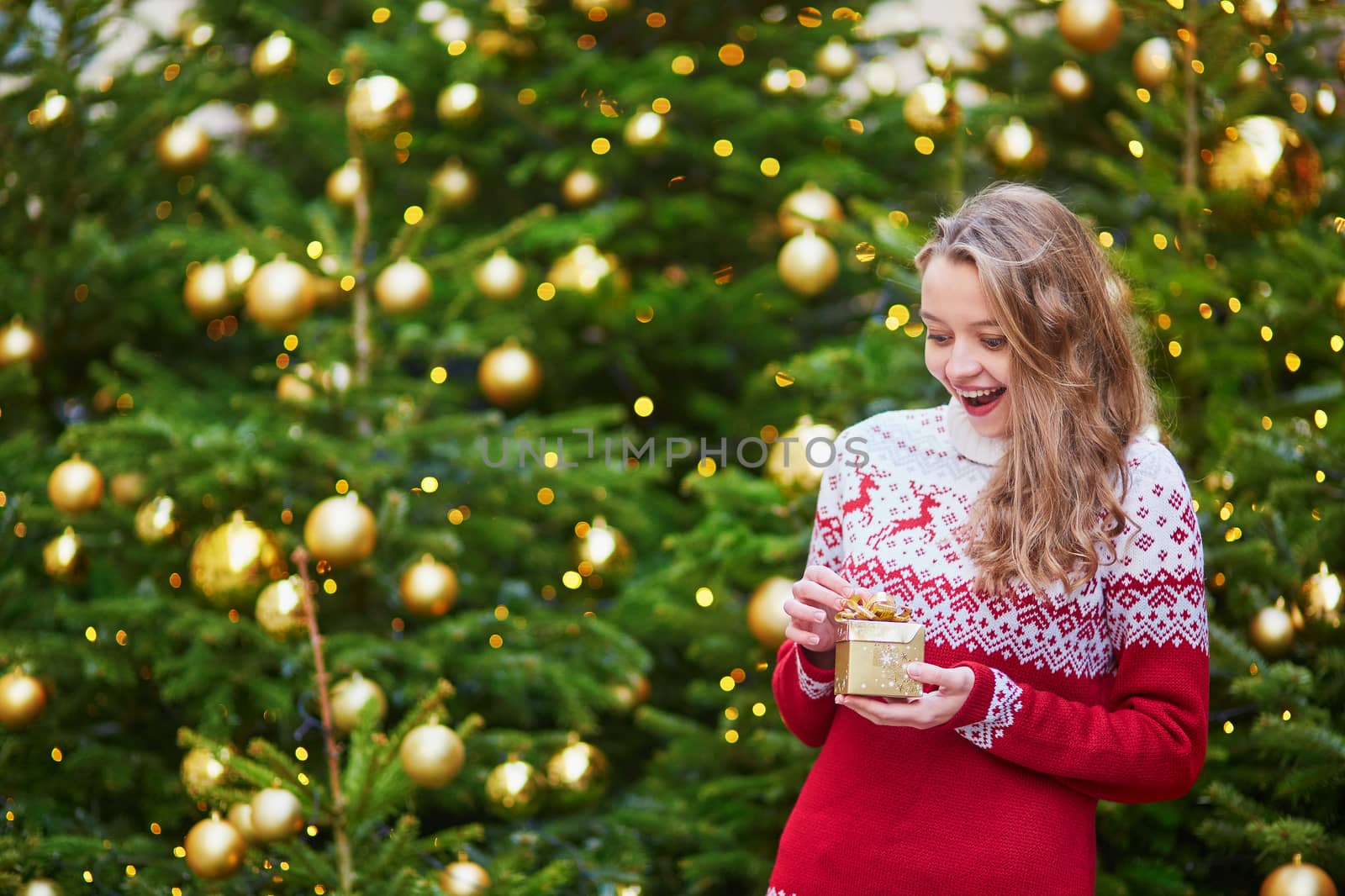 Cheerful young woman on a street of Paris decorated for Christmas, unpacking Christmas present