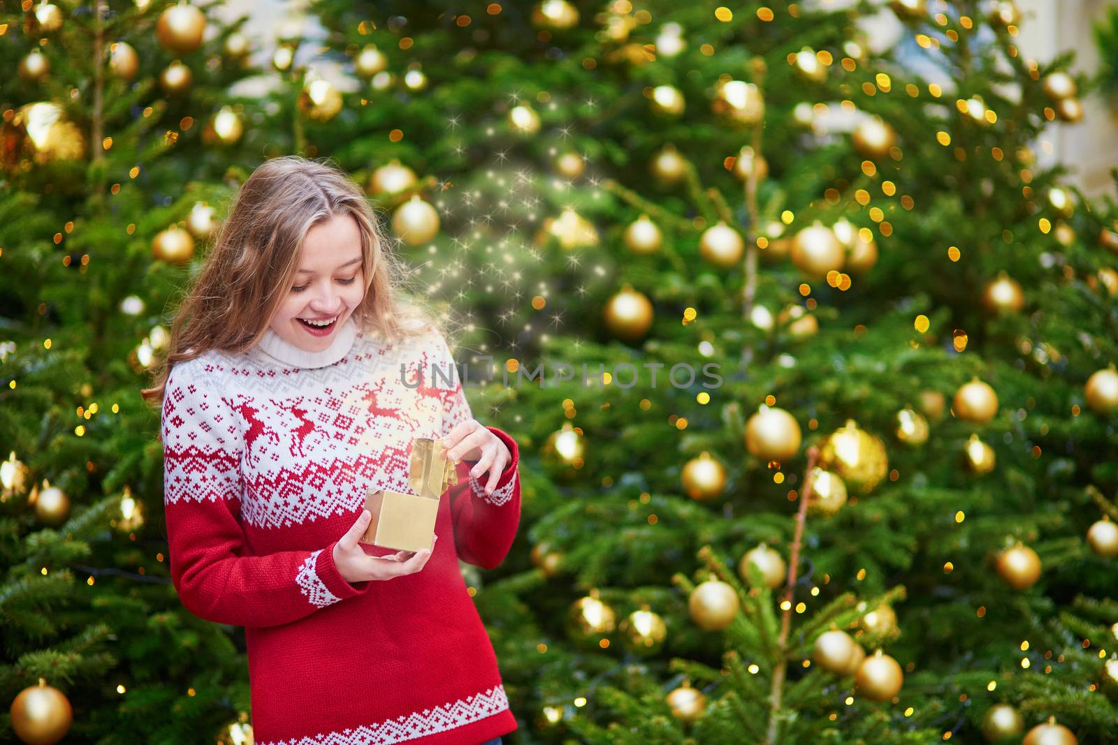 Cheerful young girl opening little present box in hands near decorated Christmas tree, magic sparkles appear from the box
