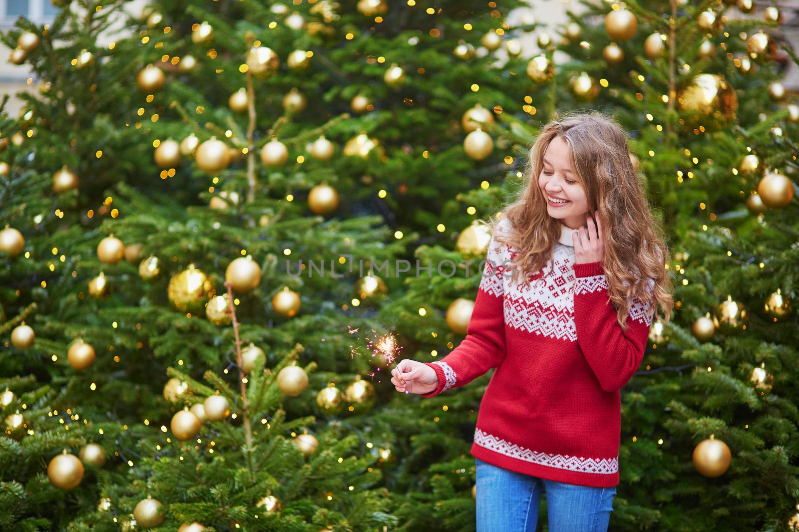 Cheerful young woman on a street of Paris decorated for Christmas with sparkling light in her hand