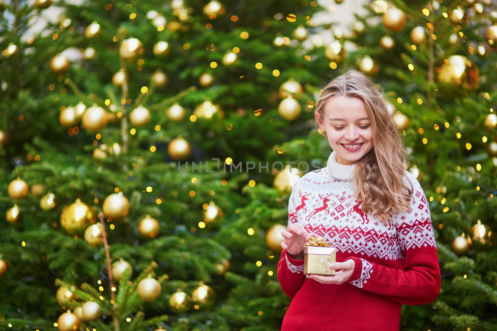 Cheerful young woman on a street of Paris decorated for Christmas, unpacking Christmas present